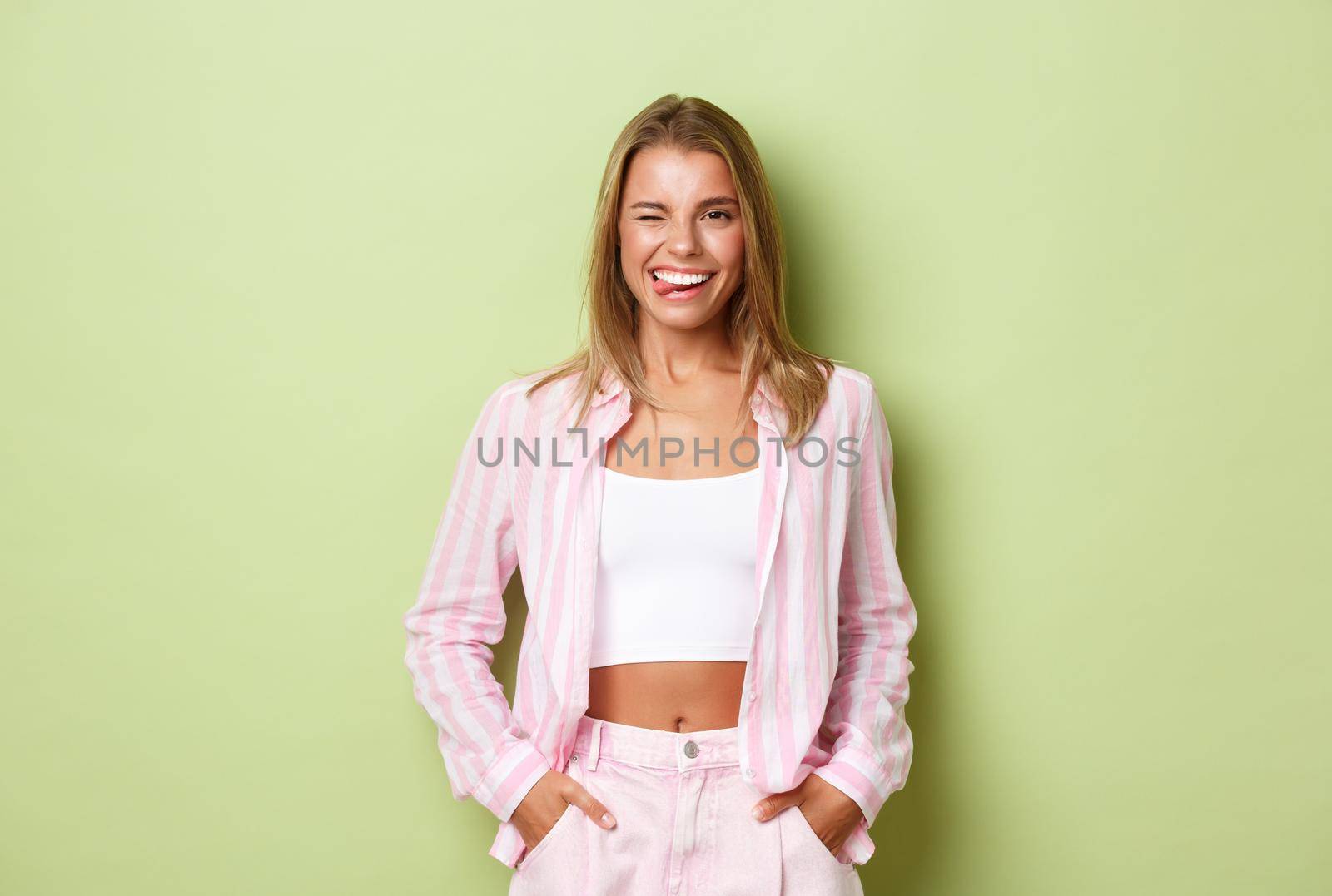 Portrait of sassy modern woman with blond short hair, wearing pink clothing, showing tongue and smiling with perfect white teeth, standing confident over green background.
