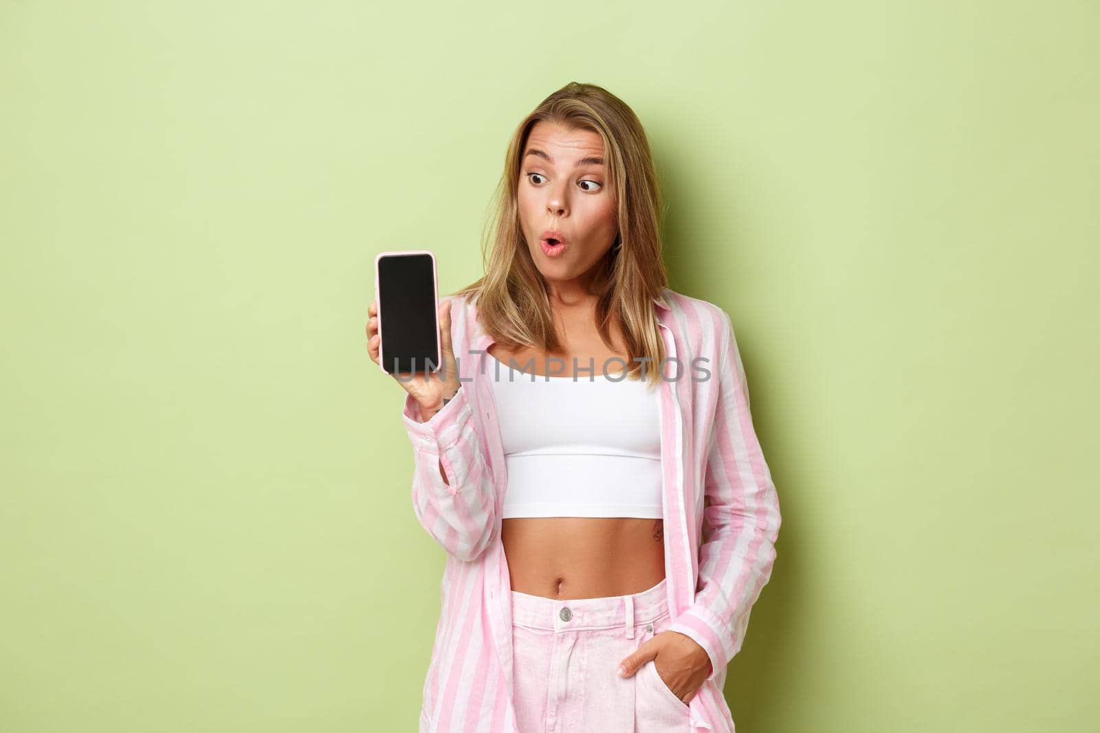 Image of blond attractive female model in pink shirt, showing smartphone screen and looking surprised, standing over green background.