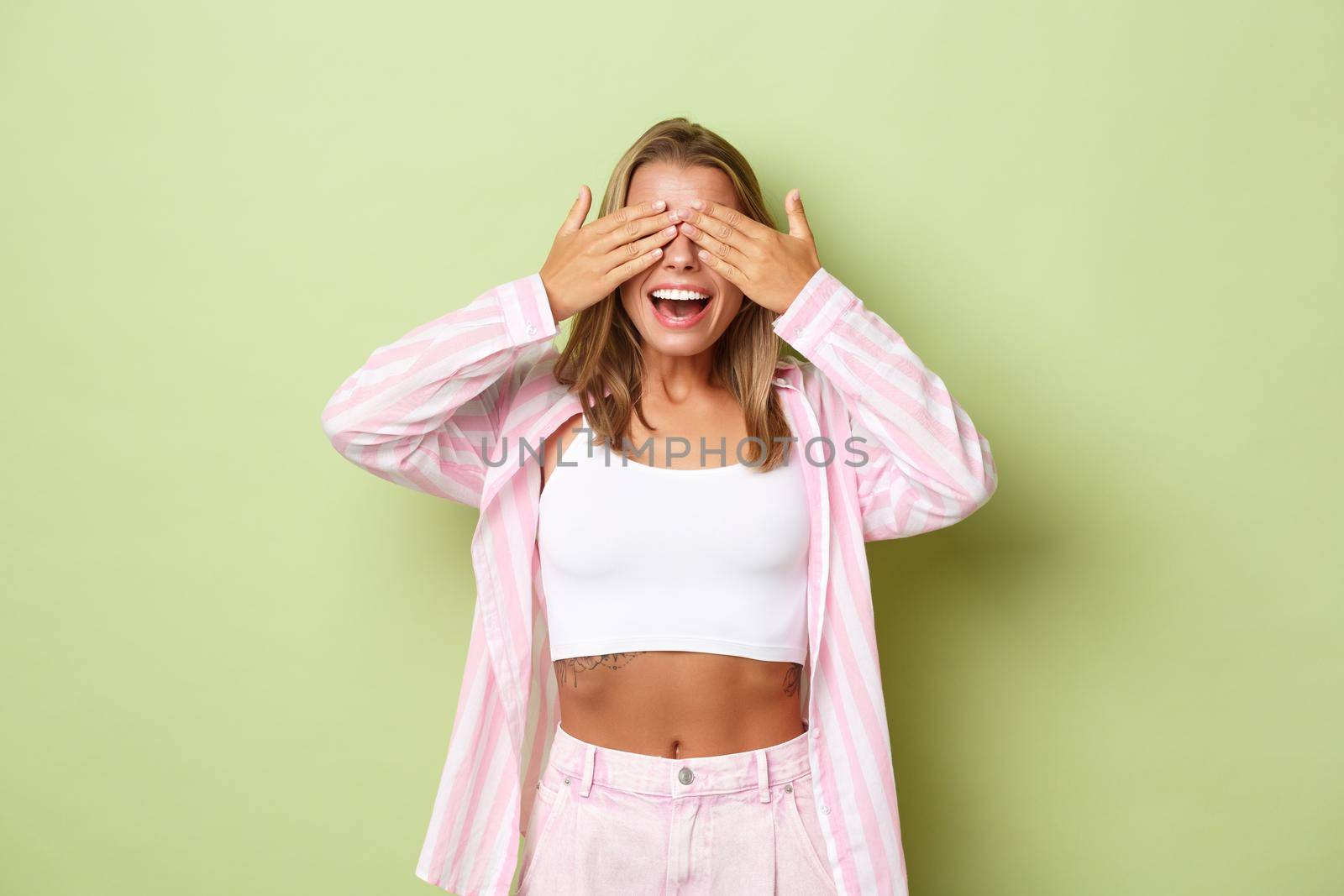 Young beautiful girl with blond short hairstyle, waiting for surprise and smiling happy, cover eyes with hands, standing over green background.