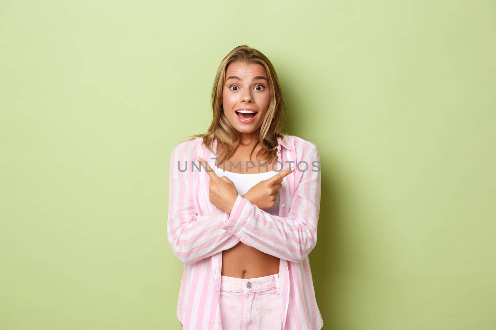 Portrait of happy smiling girl with blond short hairstyle, pointing fingers sideways with excited expression, standing in trendy pink outfit over green background by Benzoix