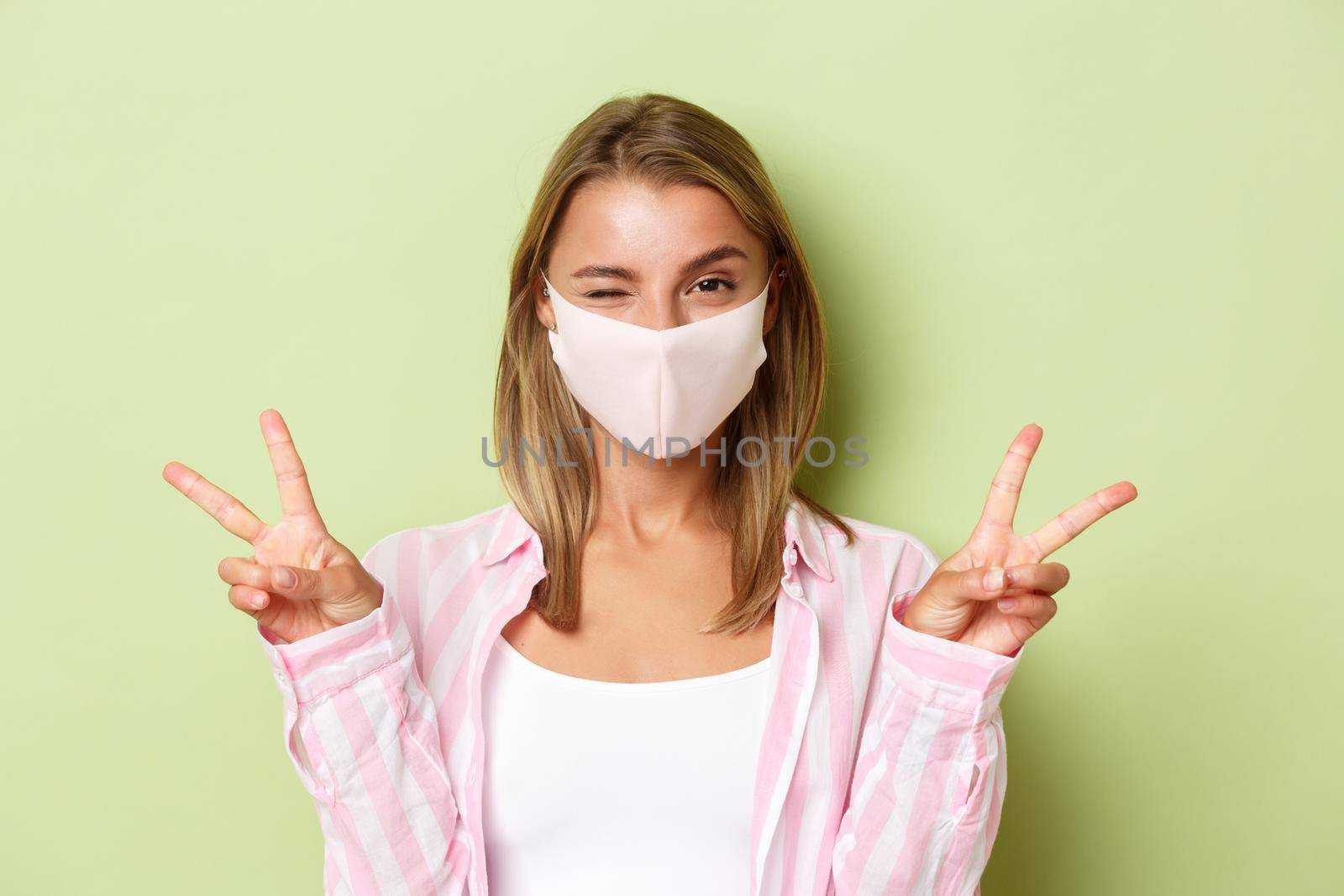 Concept of quarantine, coronavirus and lifestyle. Close-up of sassy blond girl in pink shirt and face mask, showing peace signs and winking silly, standing over green background.