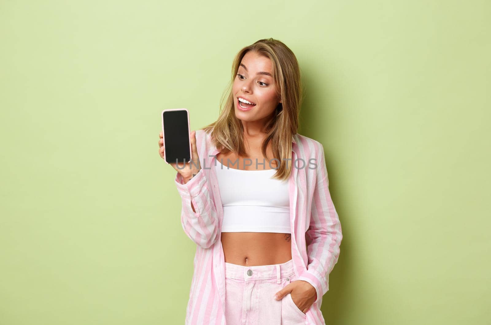 Image of blond attractive female model in pink shirt, showing smartphone screen and looking amazed, standing over green background by Benzoix