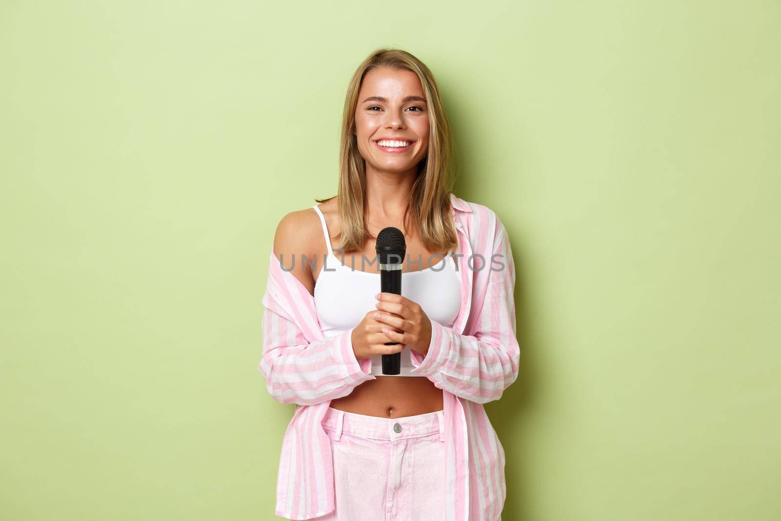 Portrait of attractive stylish woman with short blond hair, singing karaoke, holding microphone and smiling, standing over green background.