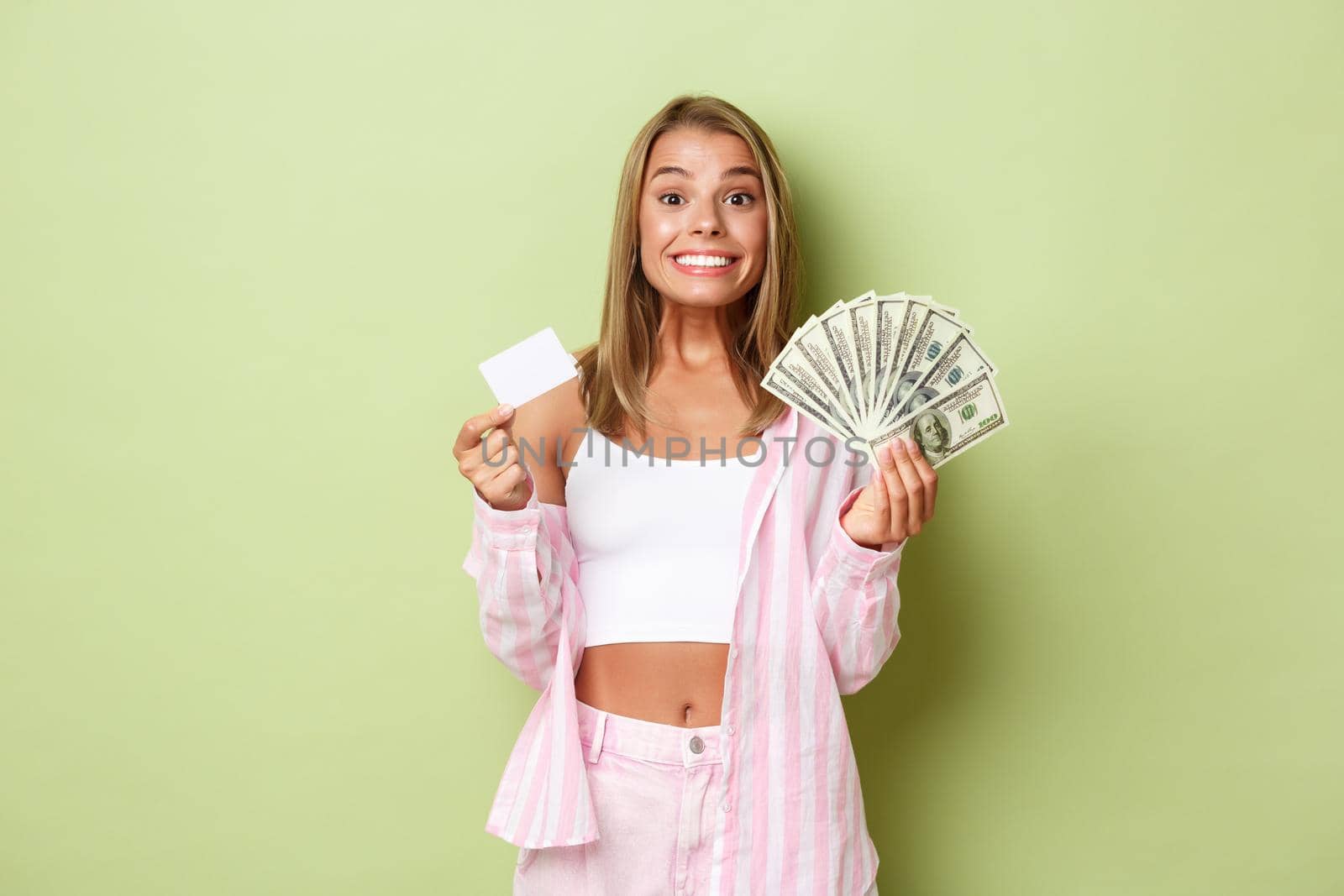 Image of successful blond woman in pink shirt, showing credit card and money, smiling happy, standing over green background.