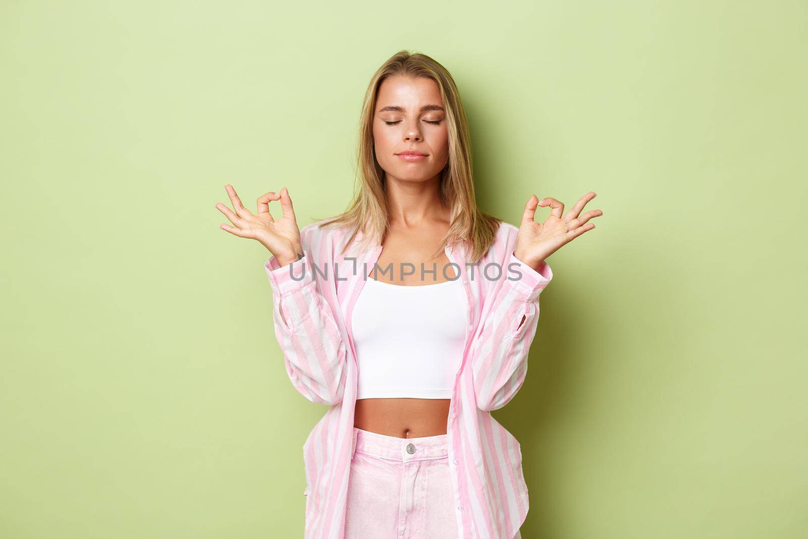 Portrait of young relaxed woman with blond short hairstyle, closing eyes and holding hands in zen gesture, meditating, practice yoga over green background.