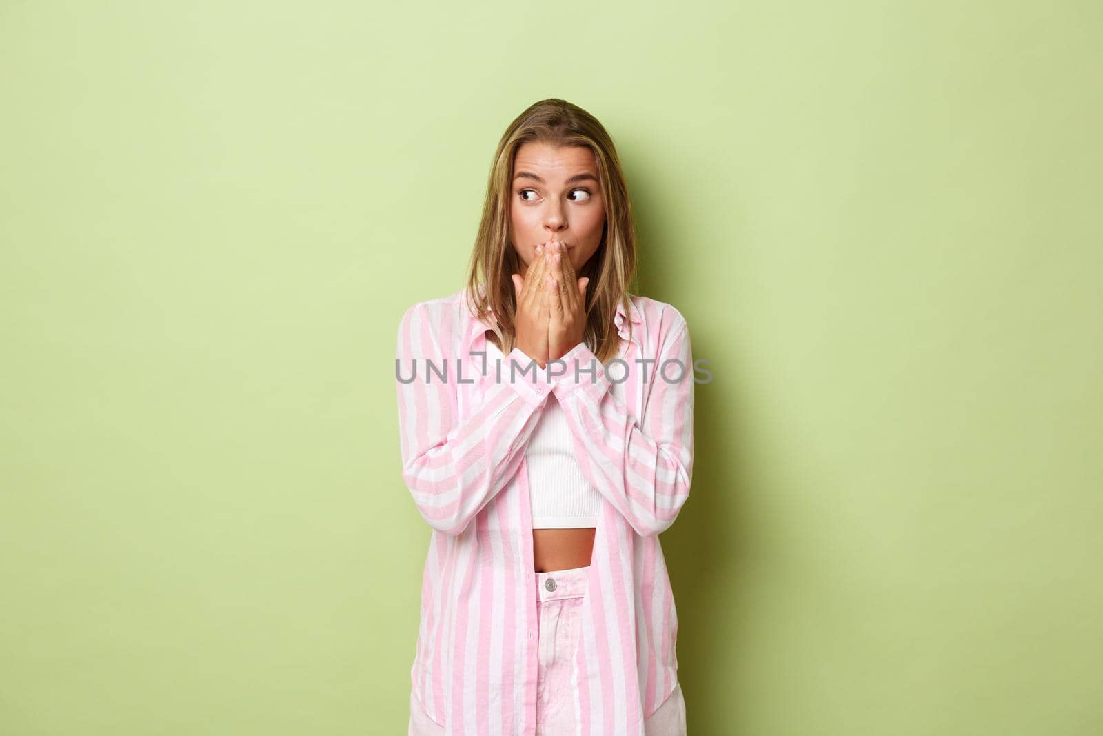 Portrait of surprised young blond girl in pink outfit, gasping and covering mouth, looking left amazed, hearing gossip, standing over green background.