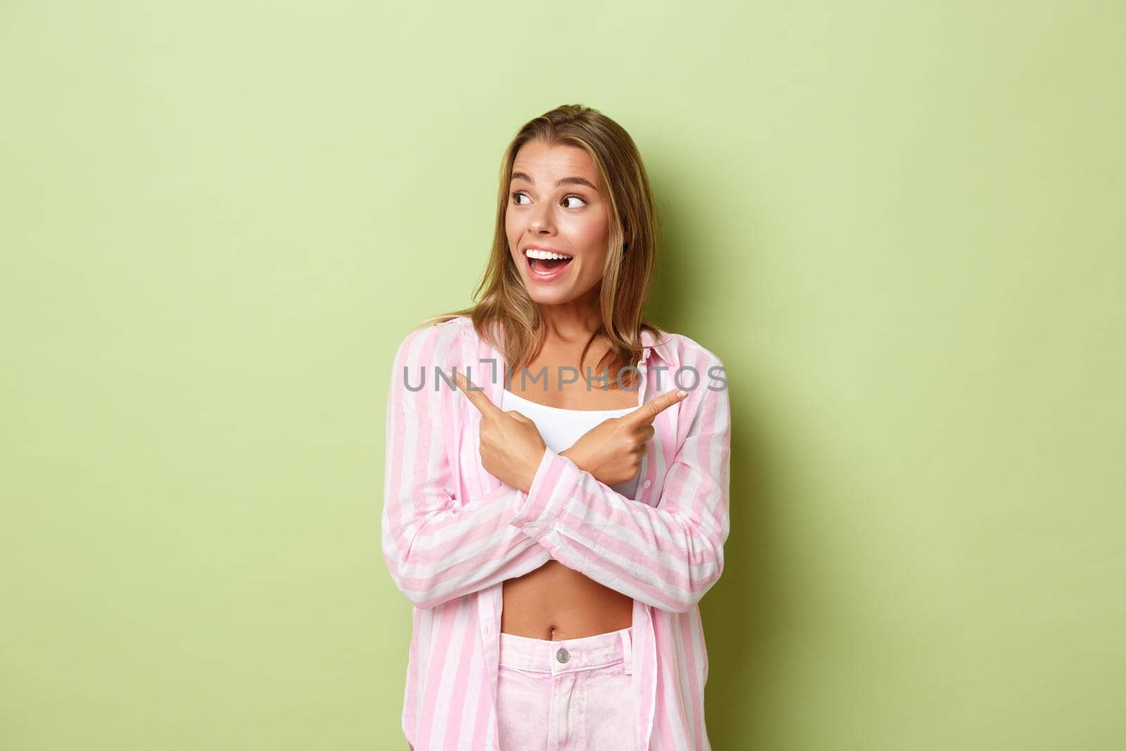 Excited attractive female model in pink outfit reacting to awesome promo, pointing fingers sideways but choosing left variant, standing over green background by Benzoix