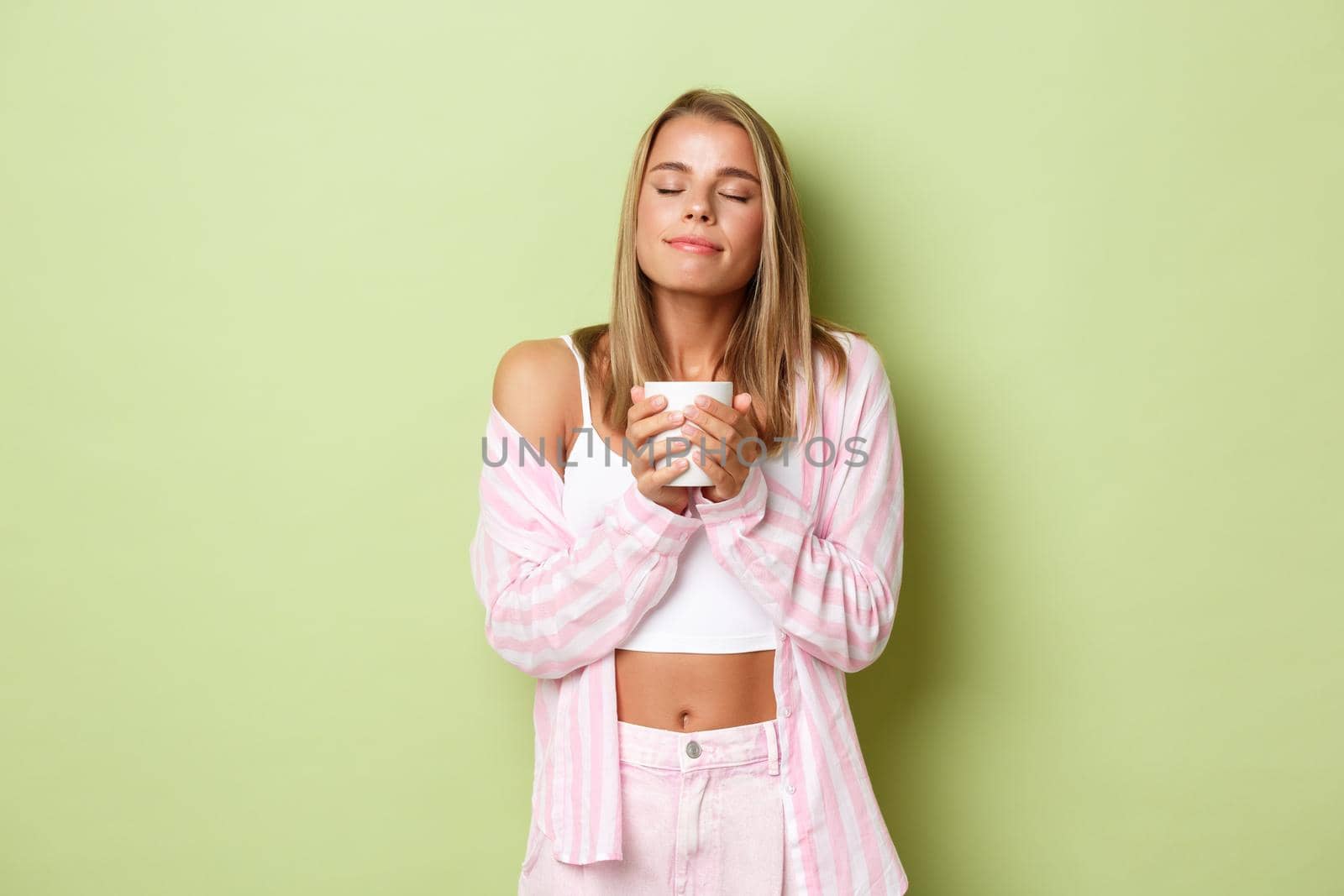 Portrait of beautiful tender woman with blond short hairstyle, smelling coffee, close eyes and smiling pleased, enjoying her drink, standing over green background.