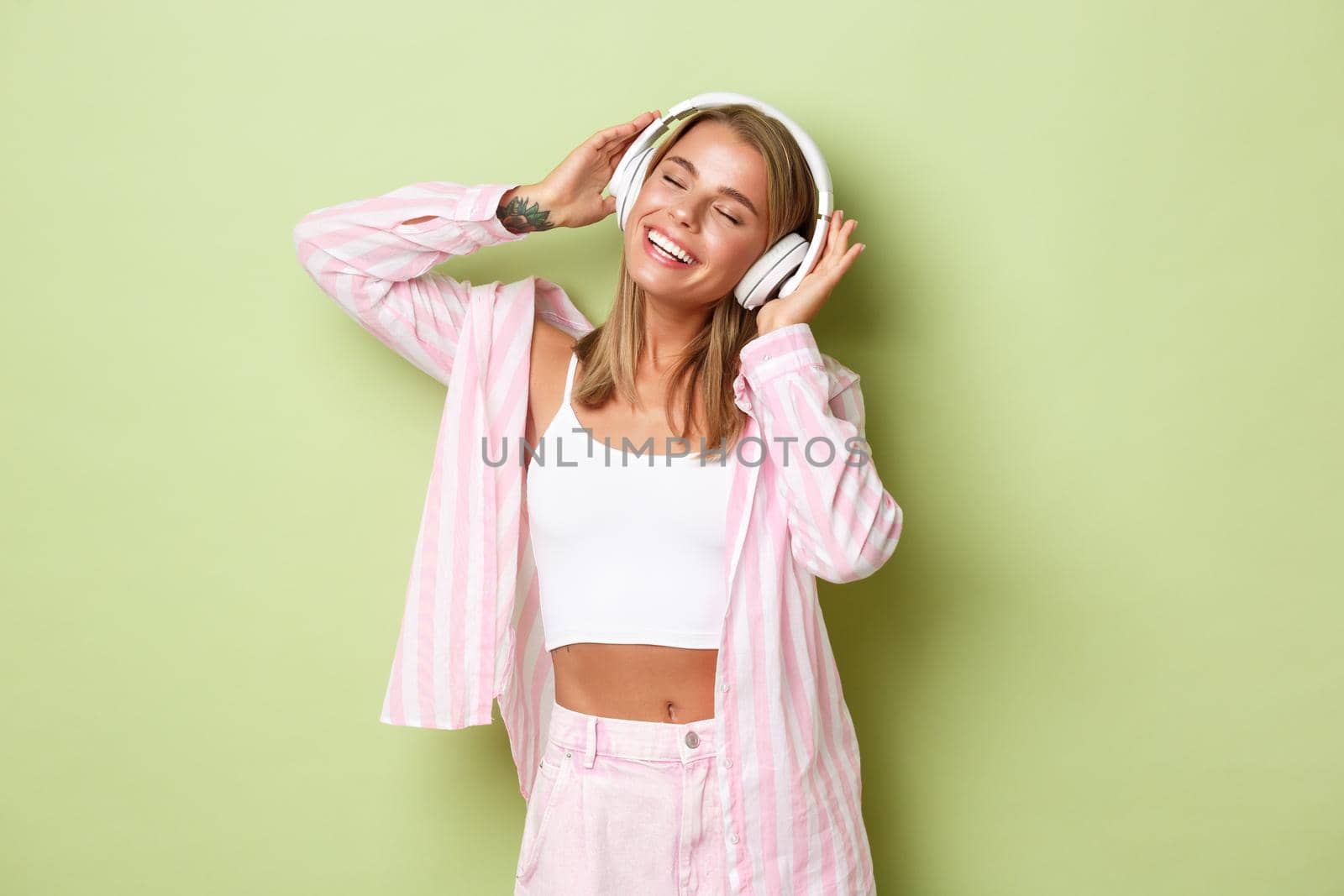 Portrait of stylish blond woman in pink shirt, with tattoos, listening to music in headphones with pleased smile, standing over green background by Benzoix