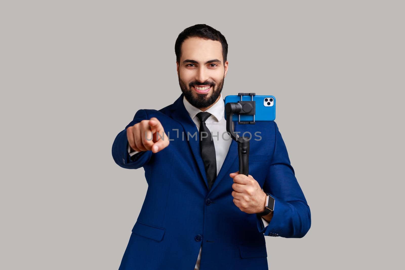 Delighted bearded man blogger holding steadicam with phone, making video or has livestream, pointing finger at camera, wearing official style suit. Indoor studio shot isolated on gray background.