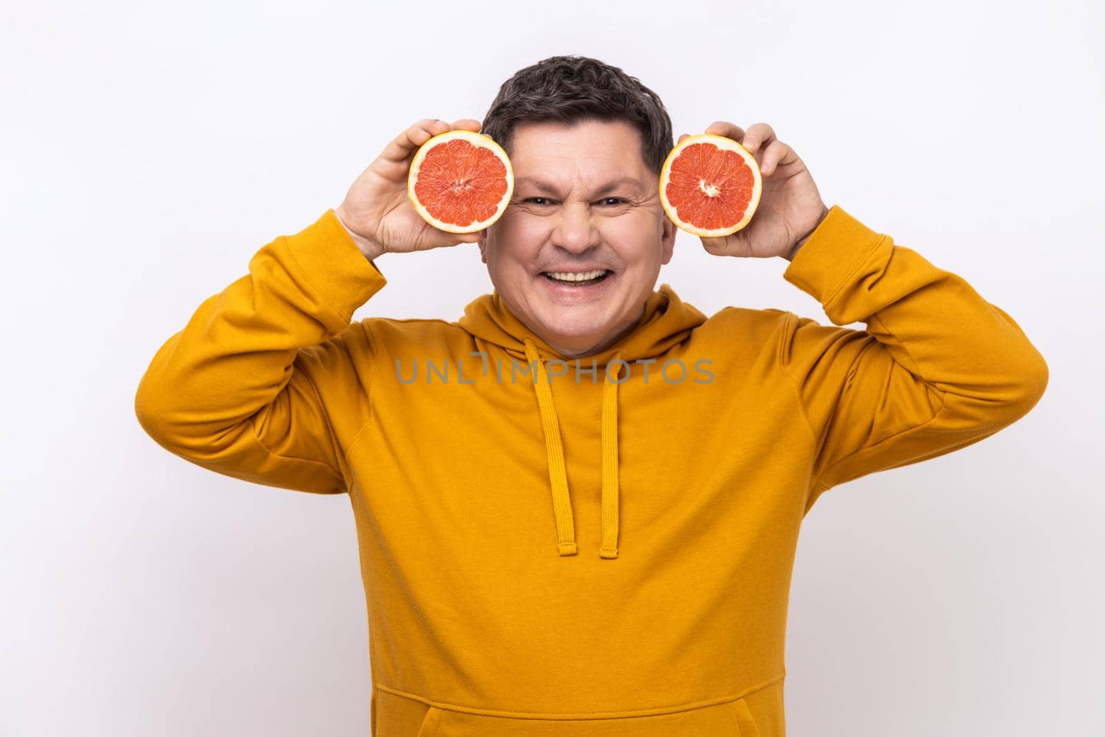 Portrait of smiling middle aged man having fun, holding two halves of grapefruit, expressing positive emotions, wearing urban style hoodie. Indoor studio shot isolated on white background.