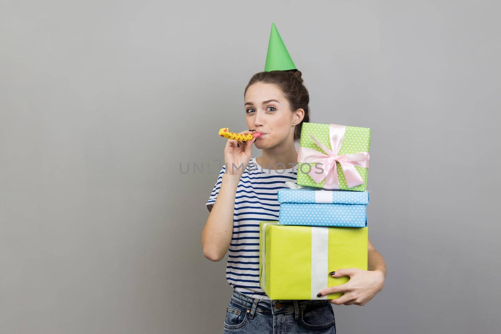 Portrait of happy delighted woman wearing striped T-shirt and party cone, holding stack of present boxes and blowing party horn. Indoor studio shot isolated on gray background.