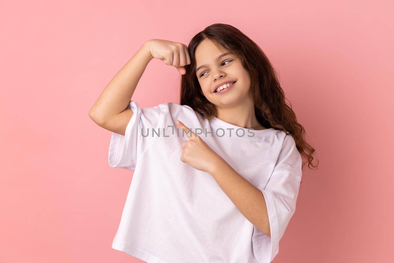 Portrait of smiling little girl wearing white T-shirt showing her arm muscle and point it with finger, proud of her strength and leadership skills. Indoor studio shot isolated on pink background.