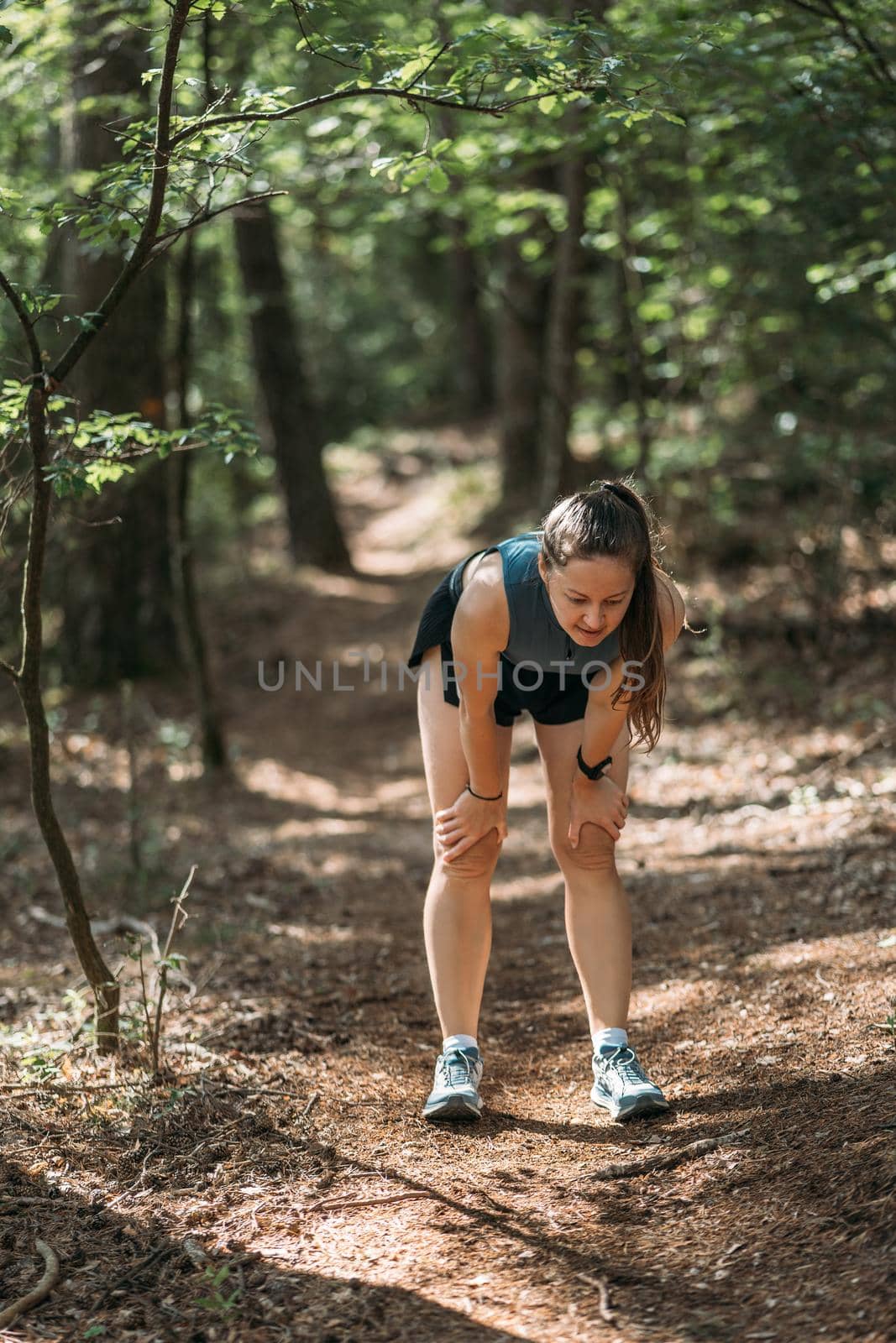Young woman in sportswear bent down and resting in the forest after working out. Trail running and active life concept