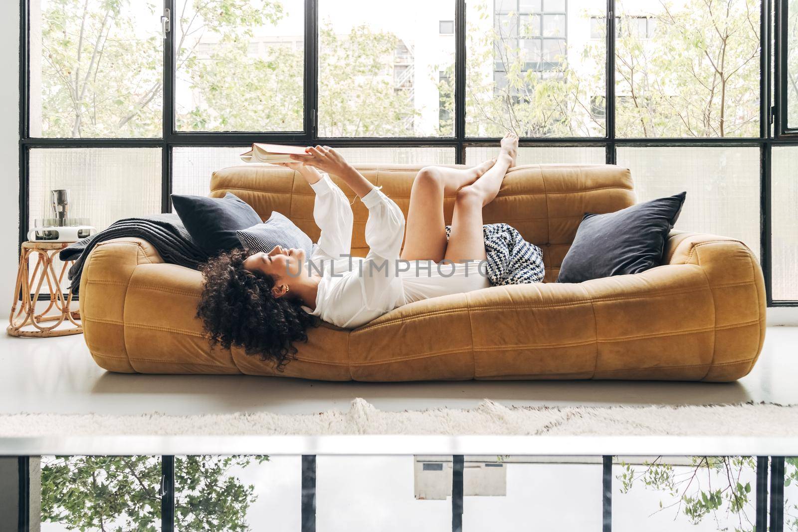 Young happy multiracial woman lying on sofa with legs up, reading a book. At home concept.