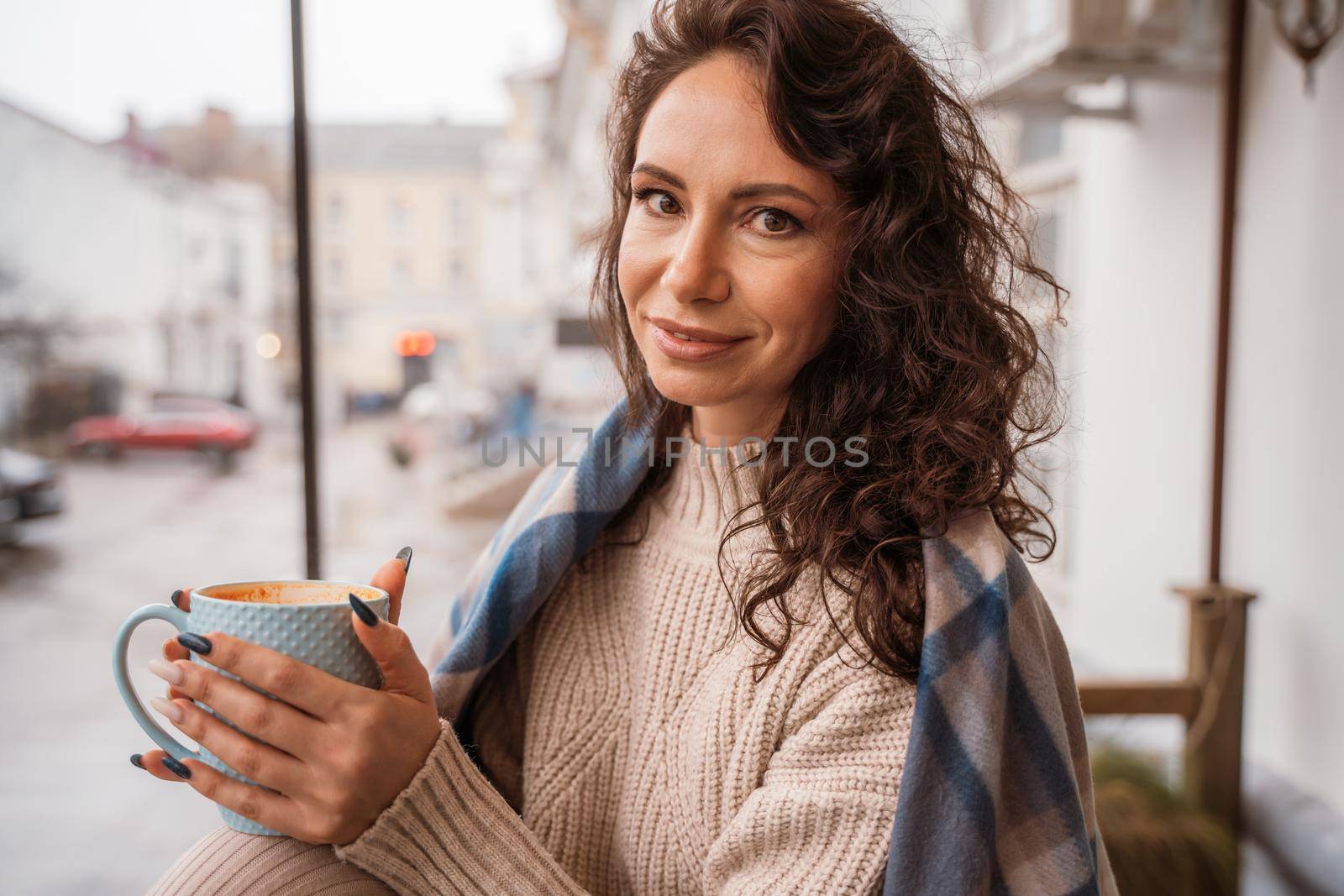 A middle-aged woman in a beige sweater with a blue mug in her hands is in a street cafe on the veranda.