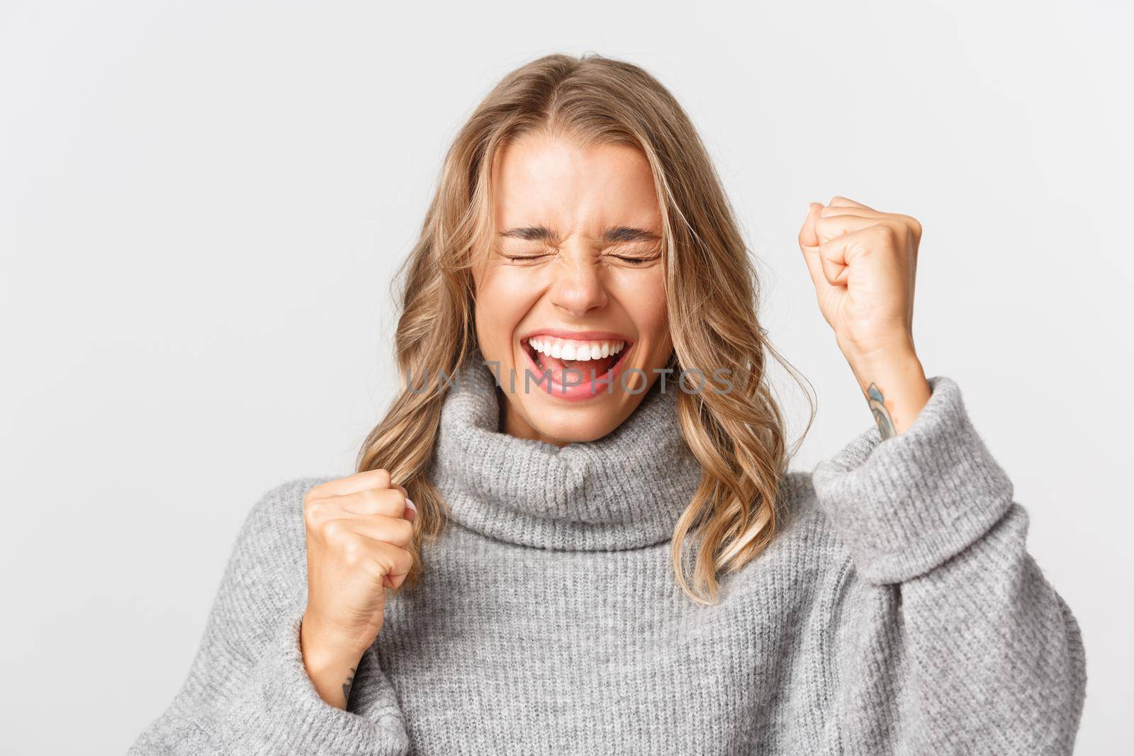 Close-up of cheerful blond girl in grey sweater, winning something and triumphing, making fist pump in rejoice, standing over white background.