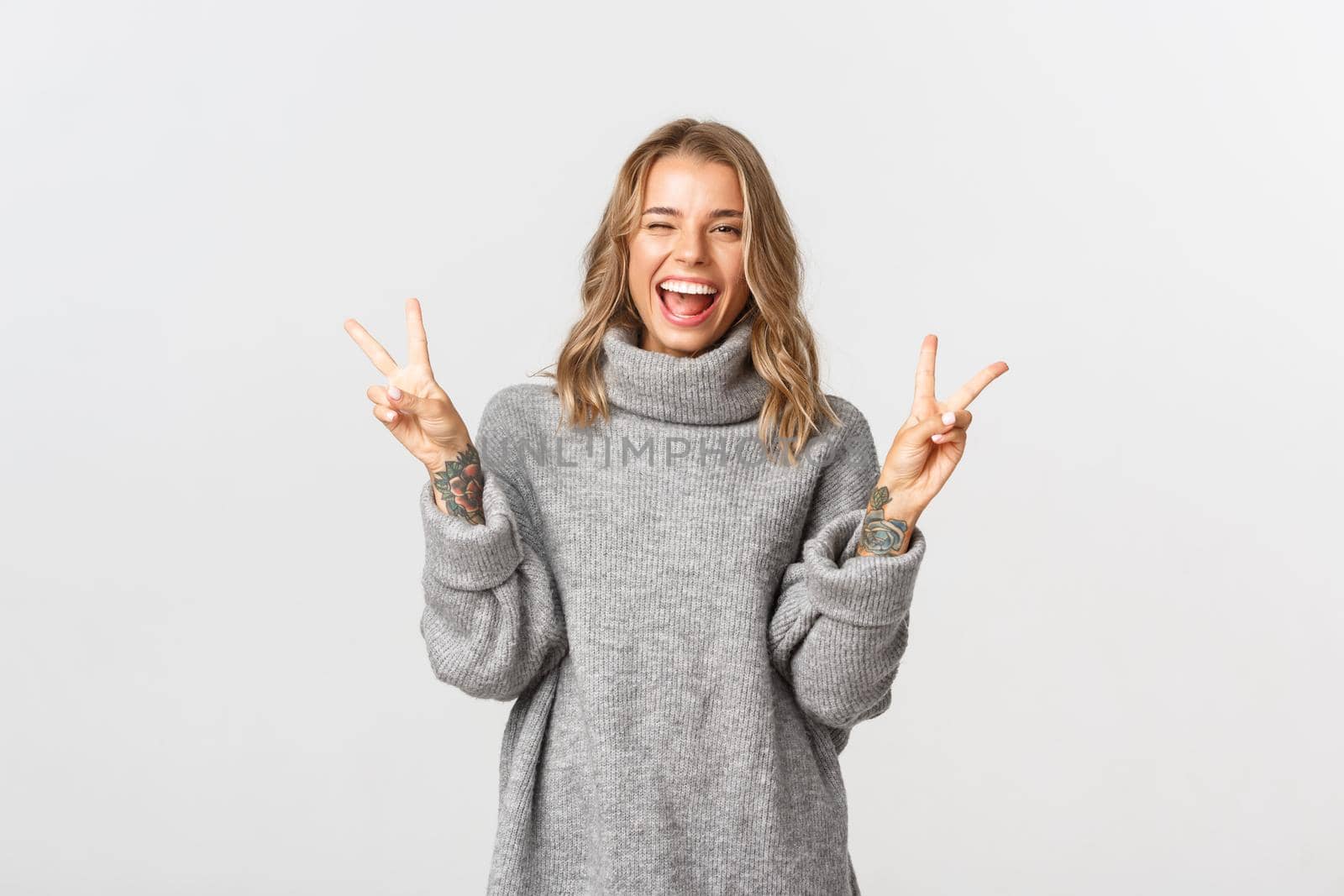 Close-up of attractive blond girl in grey sweater, showing peace signs and smiling, standing over white background.