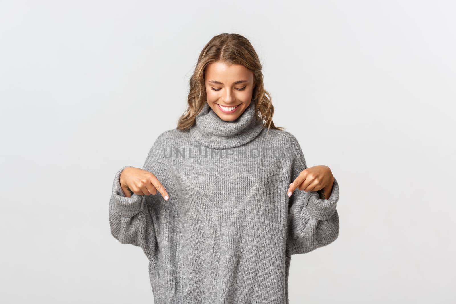 Image of lovely blond woman in grey sweater, smiling as pointing and looking down at logo advertising, standing against white background.