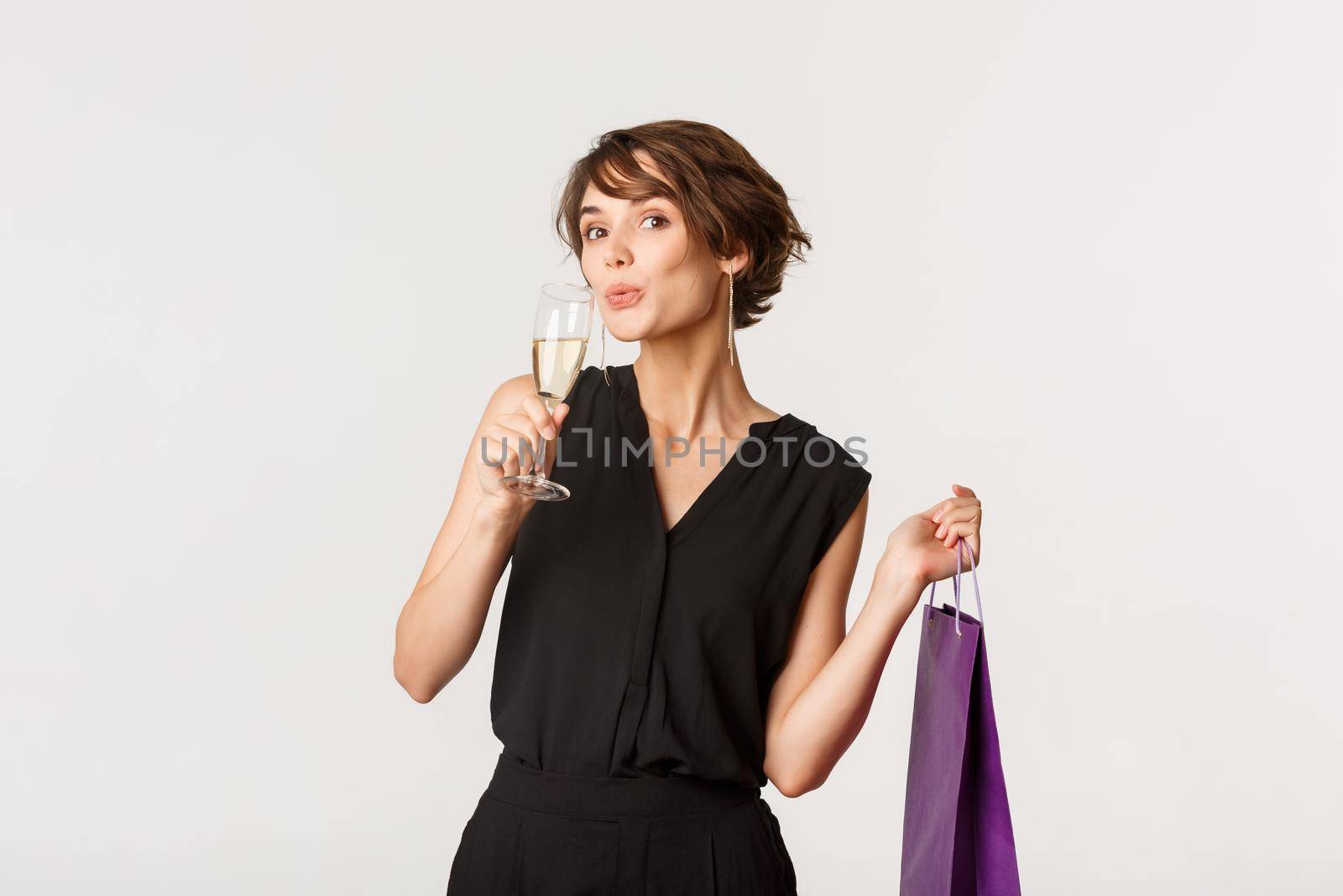 Attractive fashionable woman sipping champagne and holding gift box on formal party, standing over white background.