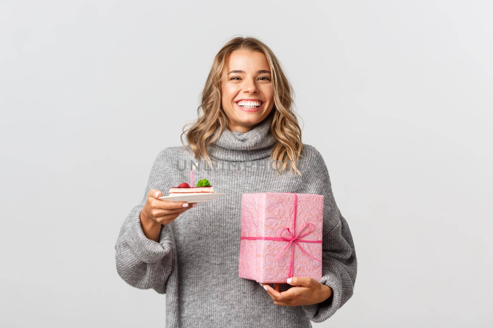 Image of happy blond woman celebrating her birthday, holding b-day cake and gift, standing over white background by Benzoix