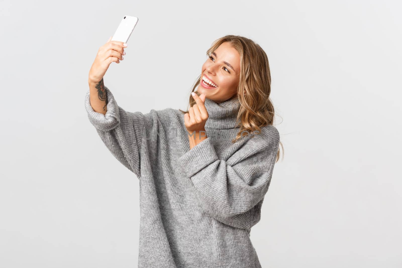 Image of happy beautiful girl in grey sweater, taking selfie on mobile phone, showing heart sign, standing over white background.