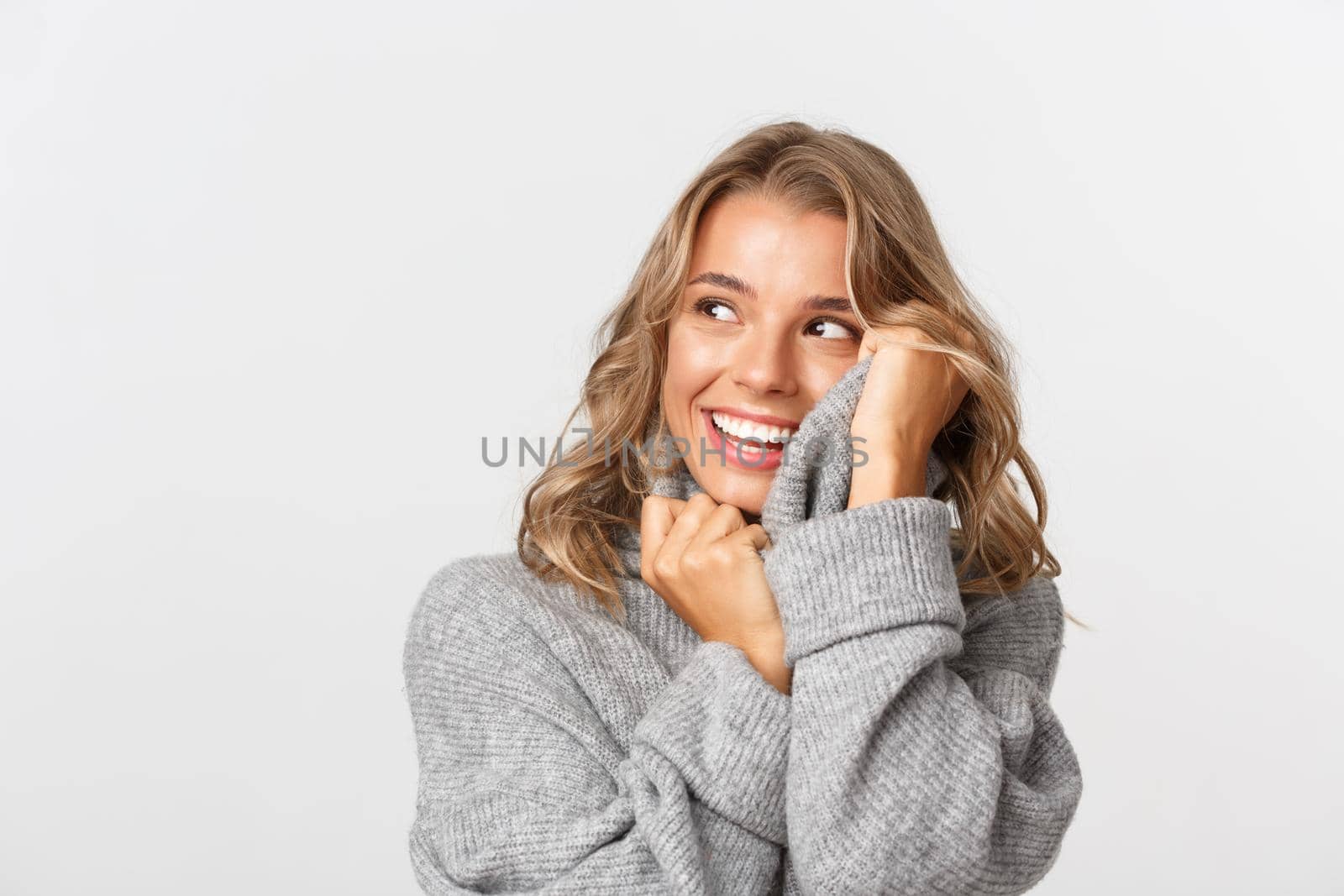 Image of attractive blond girl in grey sweater, looking happy at upper left corner, standing over white background.