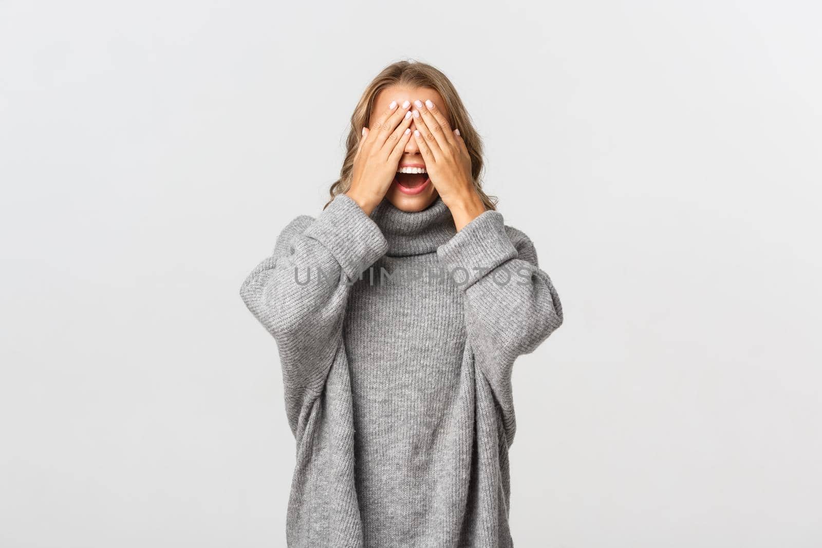 Portrait of beautiful young woman in grey sweater, standing blindsided with hands on face, waiting for surprise, standing over white background by Benzoix