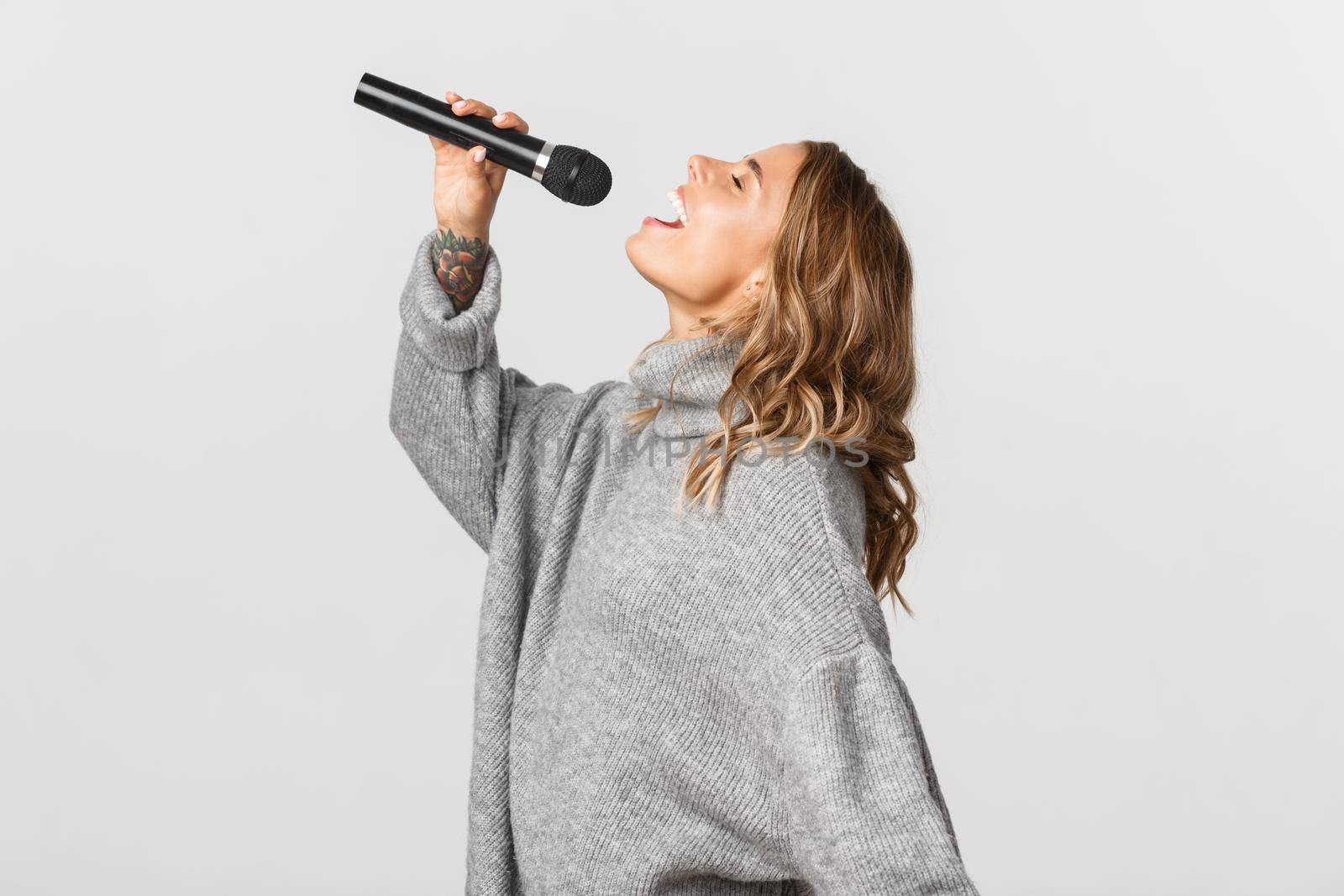 Profile shot of happy blond girl singing in microphone. Girl performing a song in karaoke, standing over white background by Benzoix