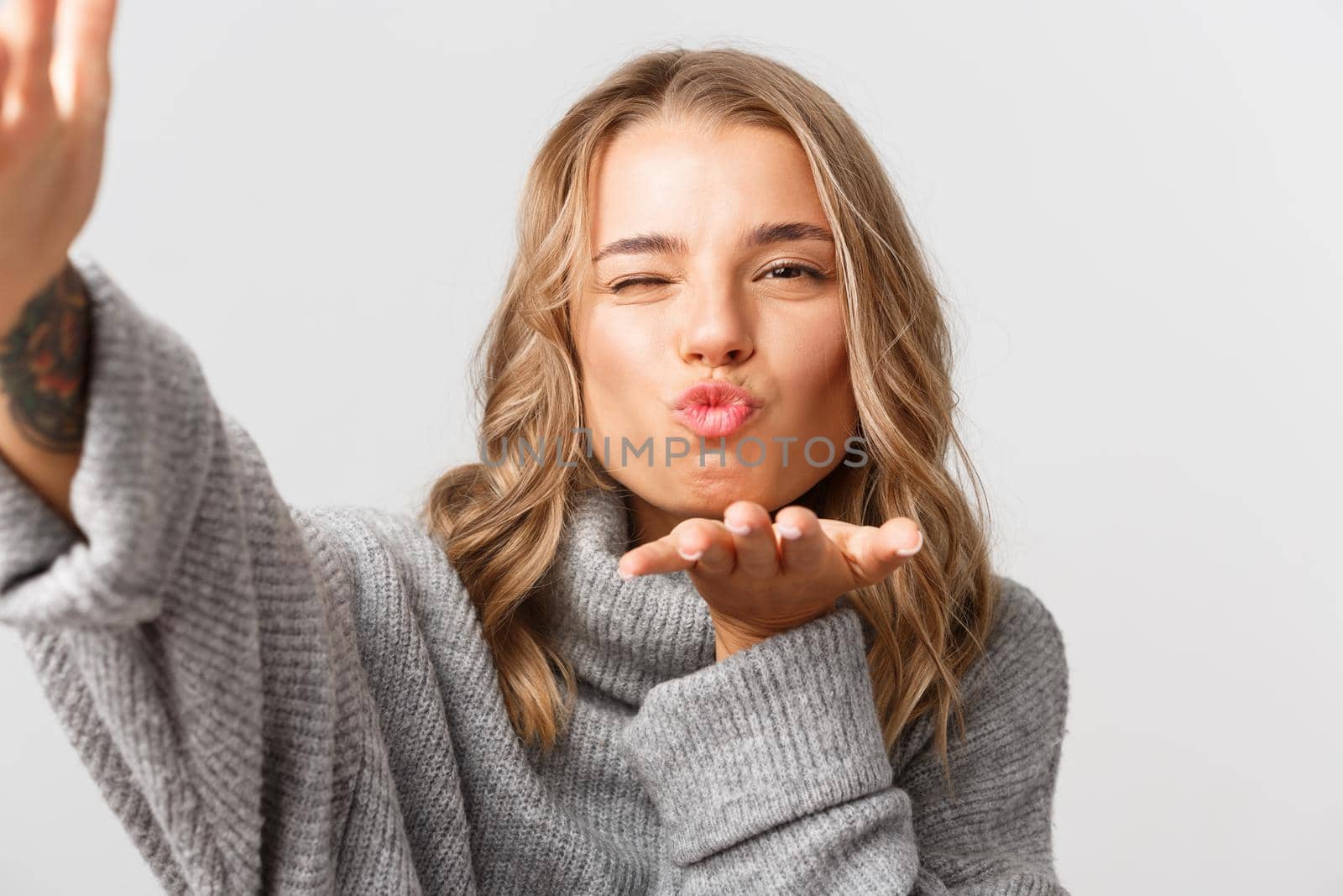 Close-up of attractive blond girl in grey sweater, taking selfie or having video call, sending air kiss at mobile camera, standing over white background by Benzoix