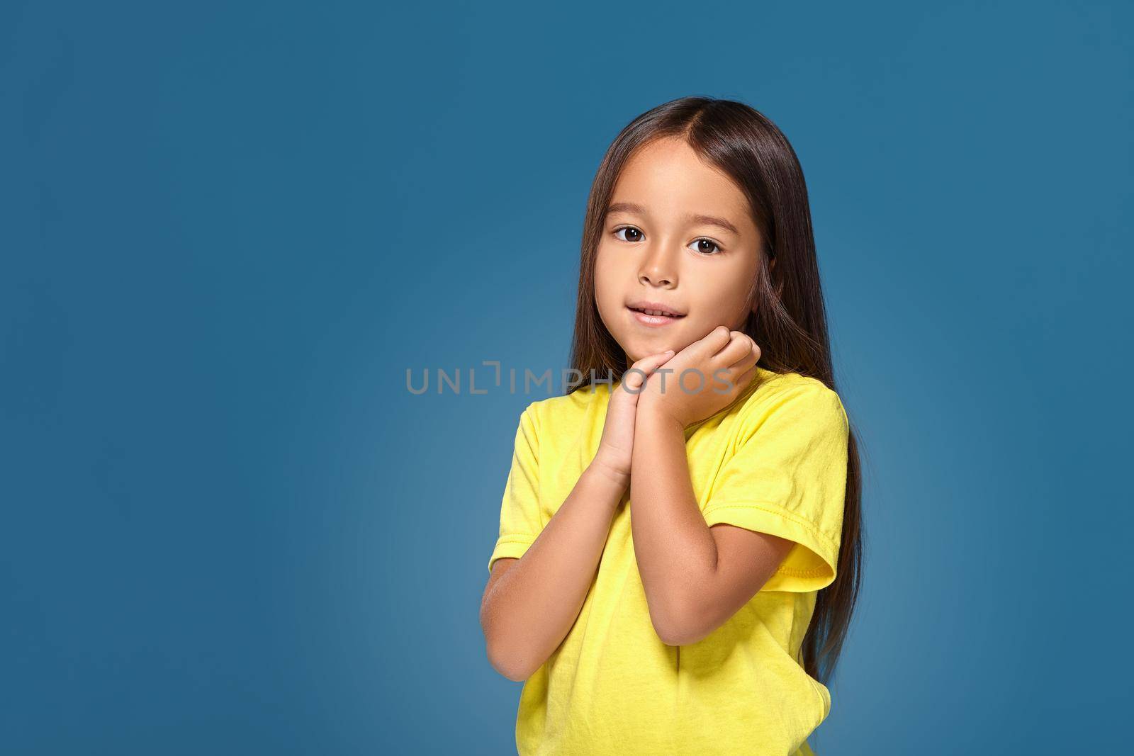 Close up portrait of cute joyful pretty littlegirl with excellent skin and beaming smile, she is admiring her beauty in a mirror, on blue background