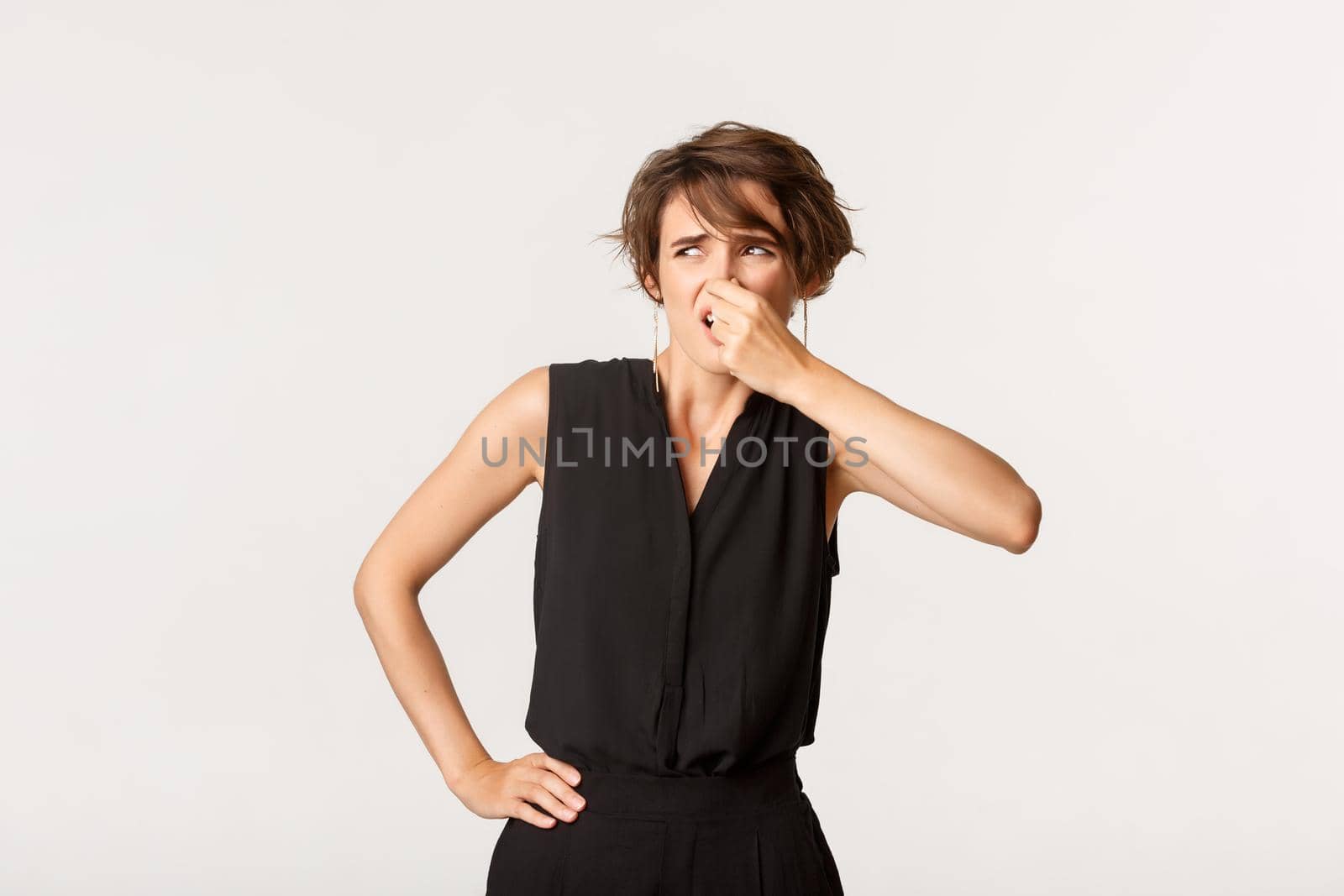 Image of young bothered woman complaining on disgusting smell, shut nose and looking left, standing white background by Benzoix