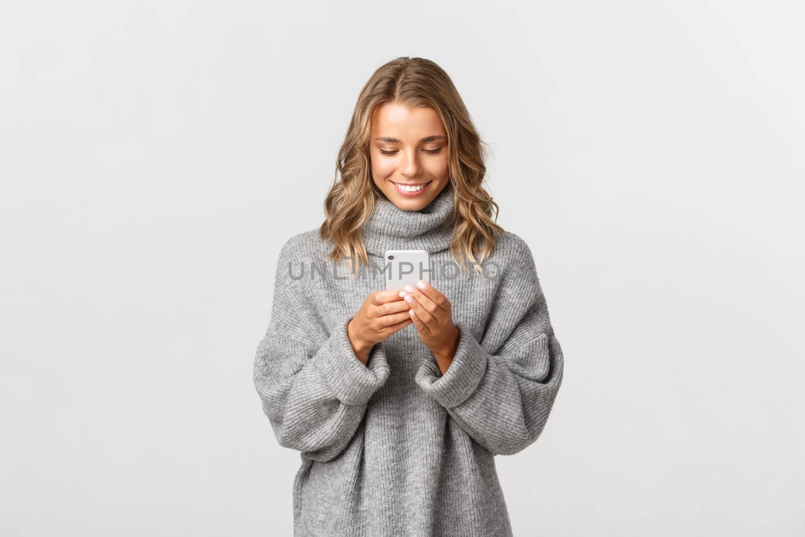 Portrait of young caucasian girl with short blond hair using mobile phone, messaging online, standing over white background.