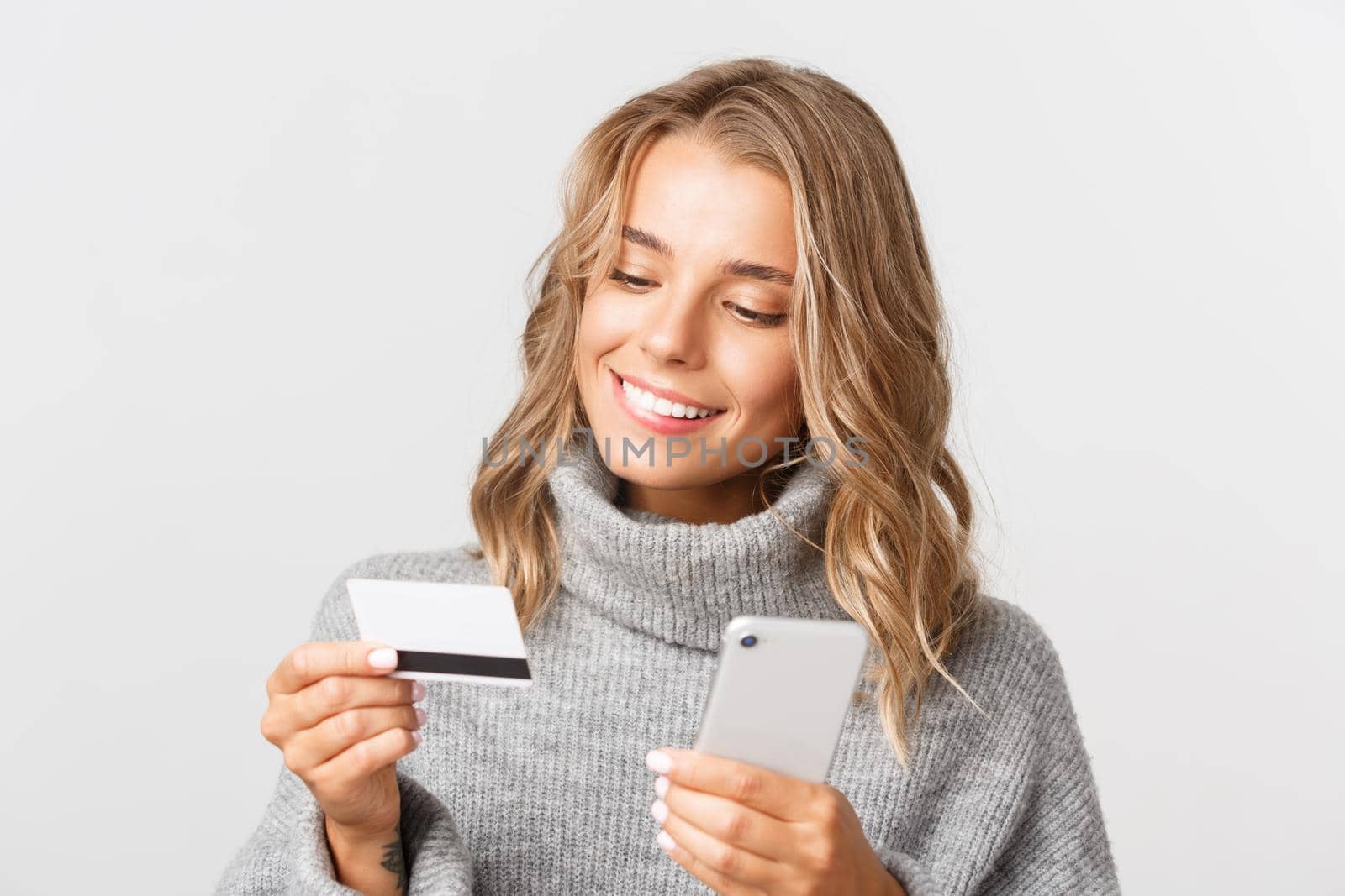 Close-up of blond woman in grey sweater looking at credit card, holding mobile phone while shopping online, standing over white background.