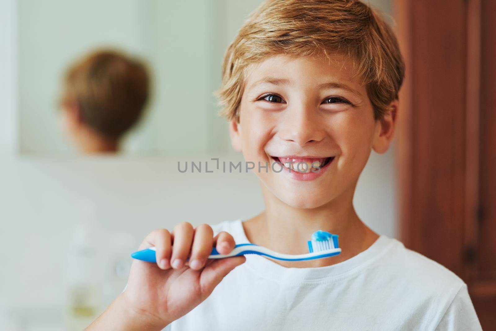 Keep your smile bright by brushing your teeth. Portrait of a cheerful young boy looking at his reflection in a mirror while brushing his teeth in the bathroom at home during the day. by YuriArcurs