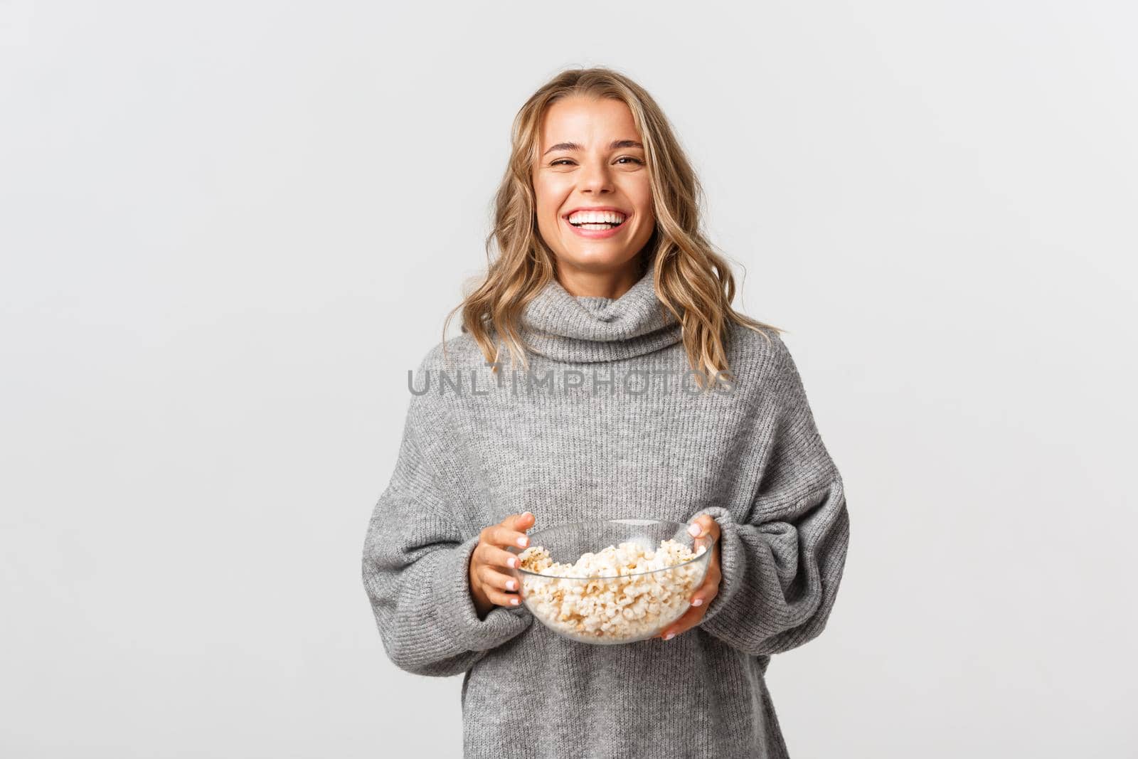 Beautiful female model with blond short hairstyle, laughing and watching tv series or movie, eating popcorn, standing over white background.