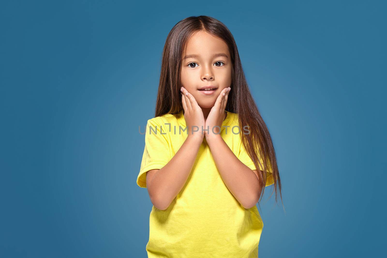 Close up portrait of cute joyful pretty littlegirl with excellent skin and beaming smile, she is admiring her beauty in a mirror, on blue background