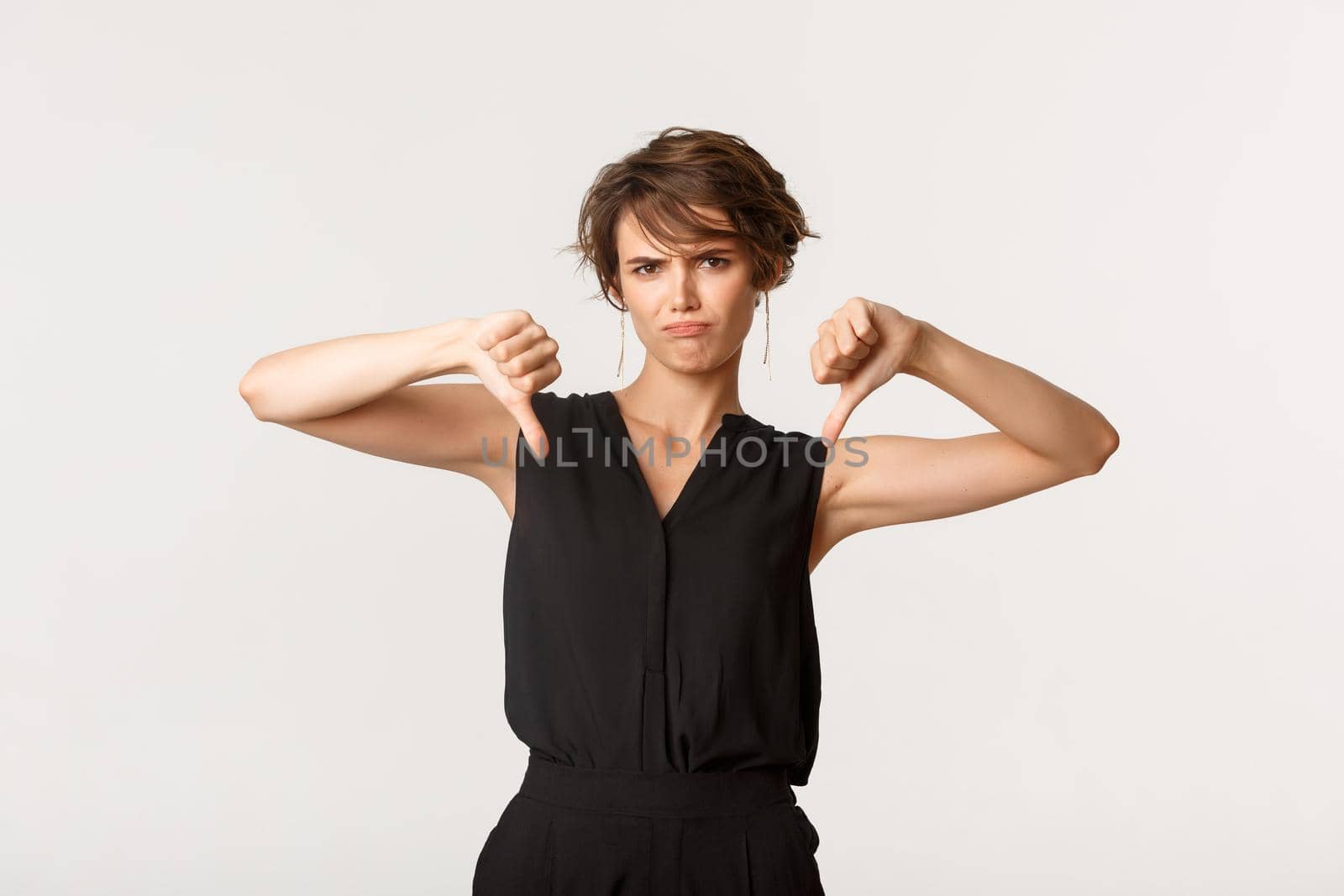 Disappointed young woman showing thumbs-down and grimacing judgemental, dislike product, leave negative feedback, standing white background.