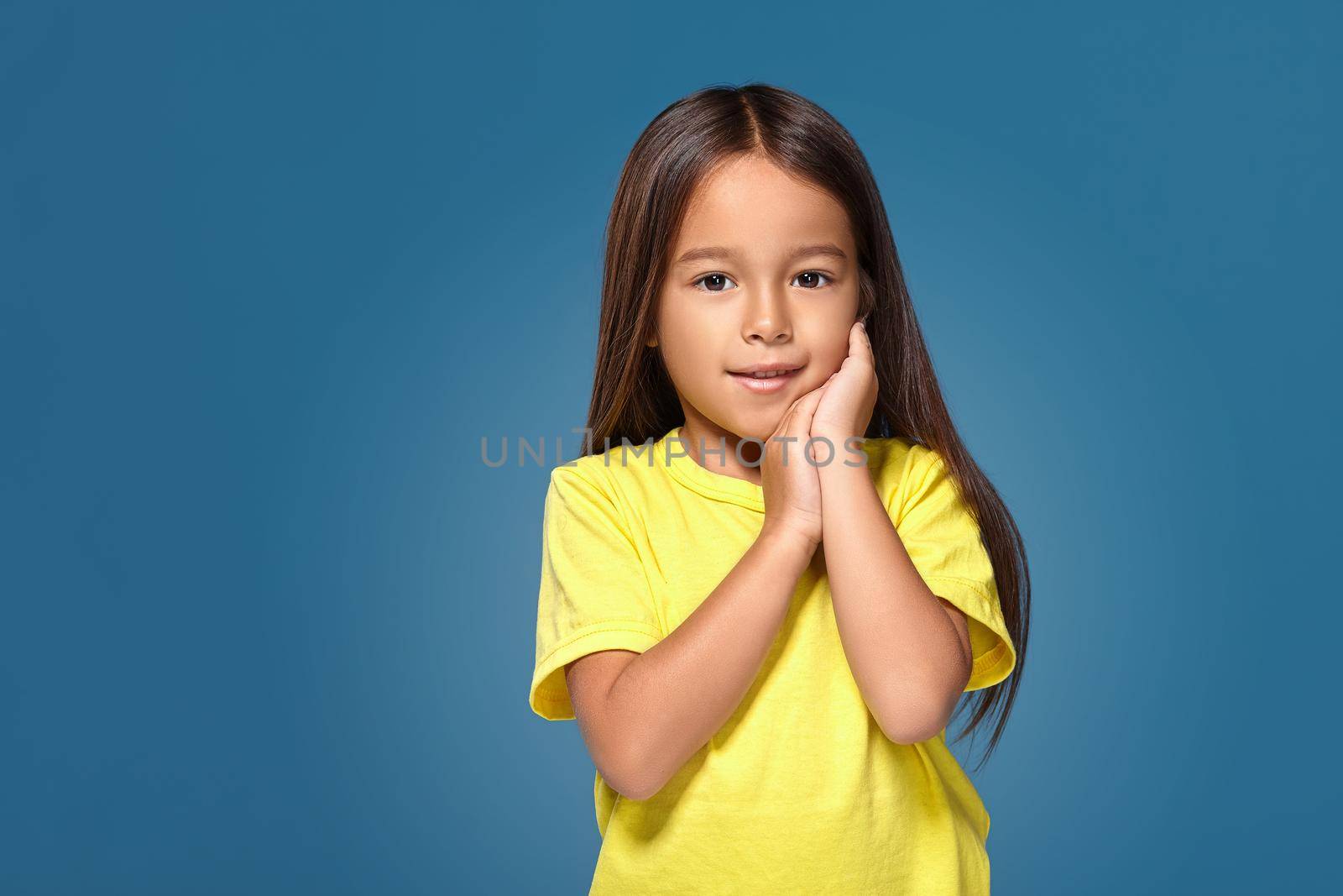 Close up portrait of cute joyful pretty littlegirl with excellent skin and beaming smile, she is admiring her beauty in a mirror, on blue background