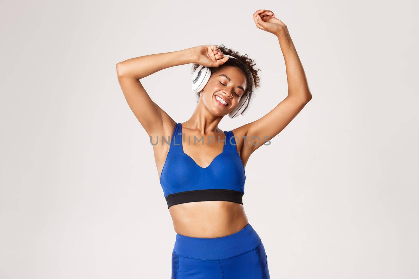 Sport and lifestyle concept. Happy fit african-american woman dancing, smiling with eyes closed, listening music in wireless headphones during workout, white background.
