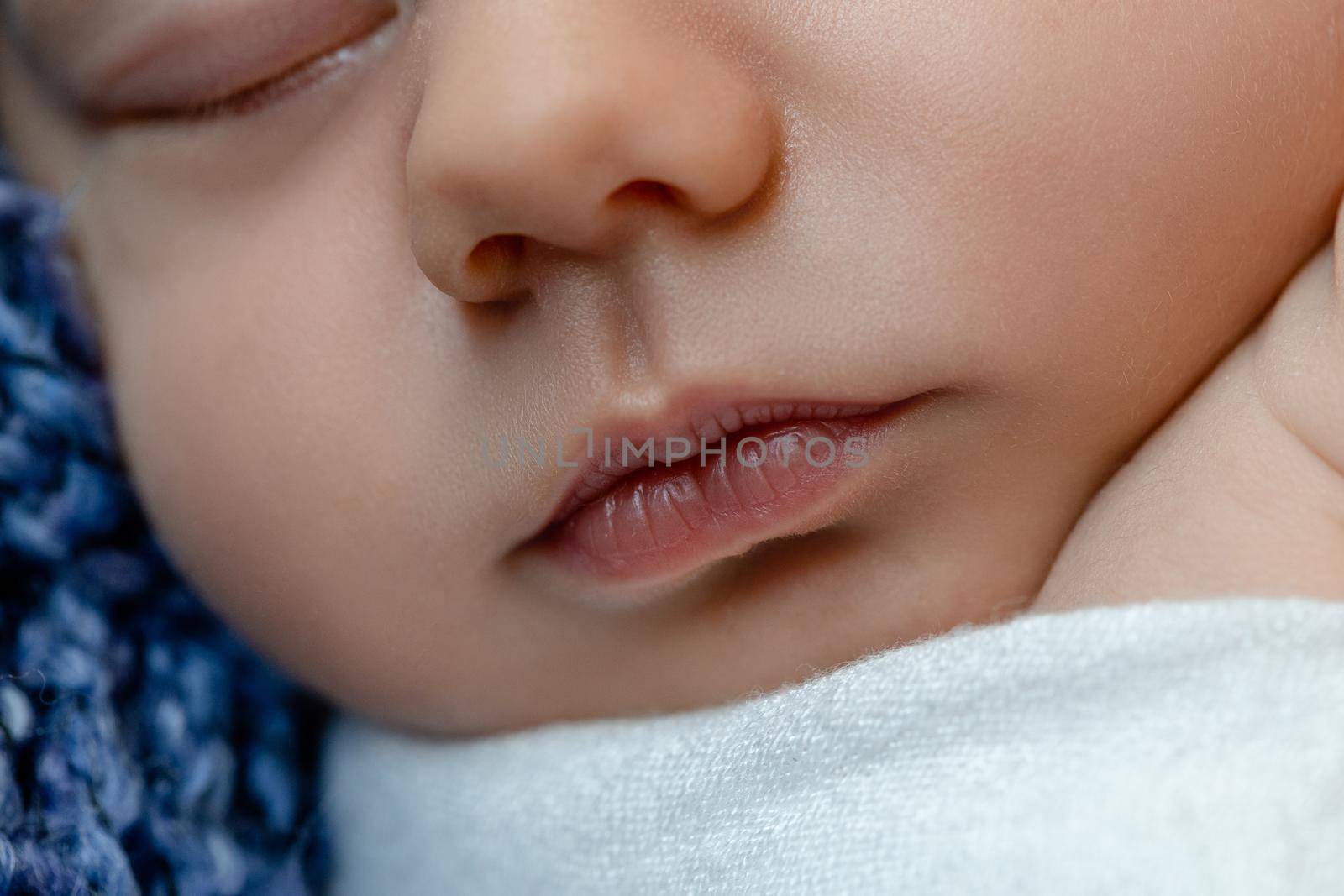 Newborn - baby, face close-up. The sleeping Newborn boy under a white knitted blanket lies on the blue fur. Newborn. 14 days.