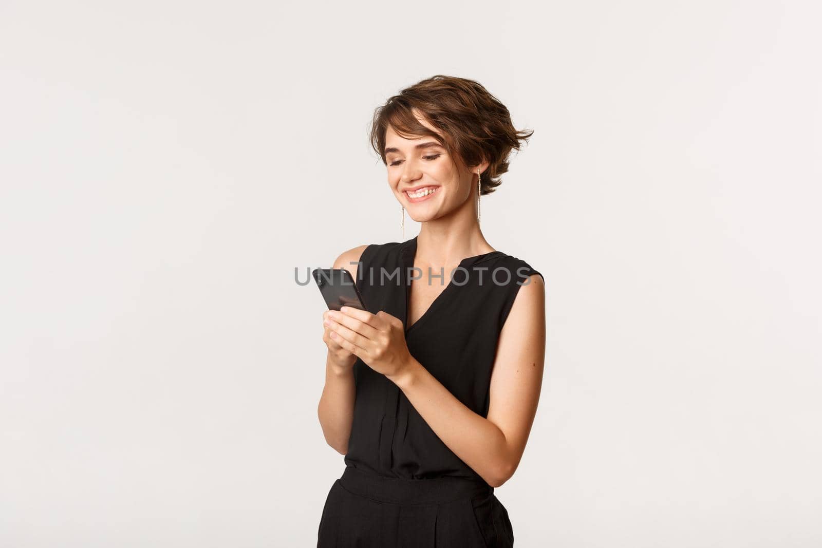 Portrait of successful young woman holding smartphone and smiling, chatting or browsing social networks, standing white background.