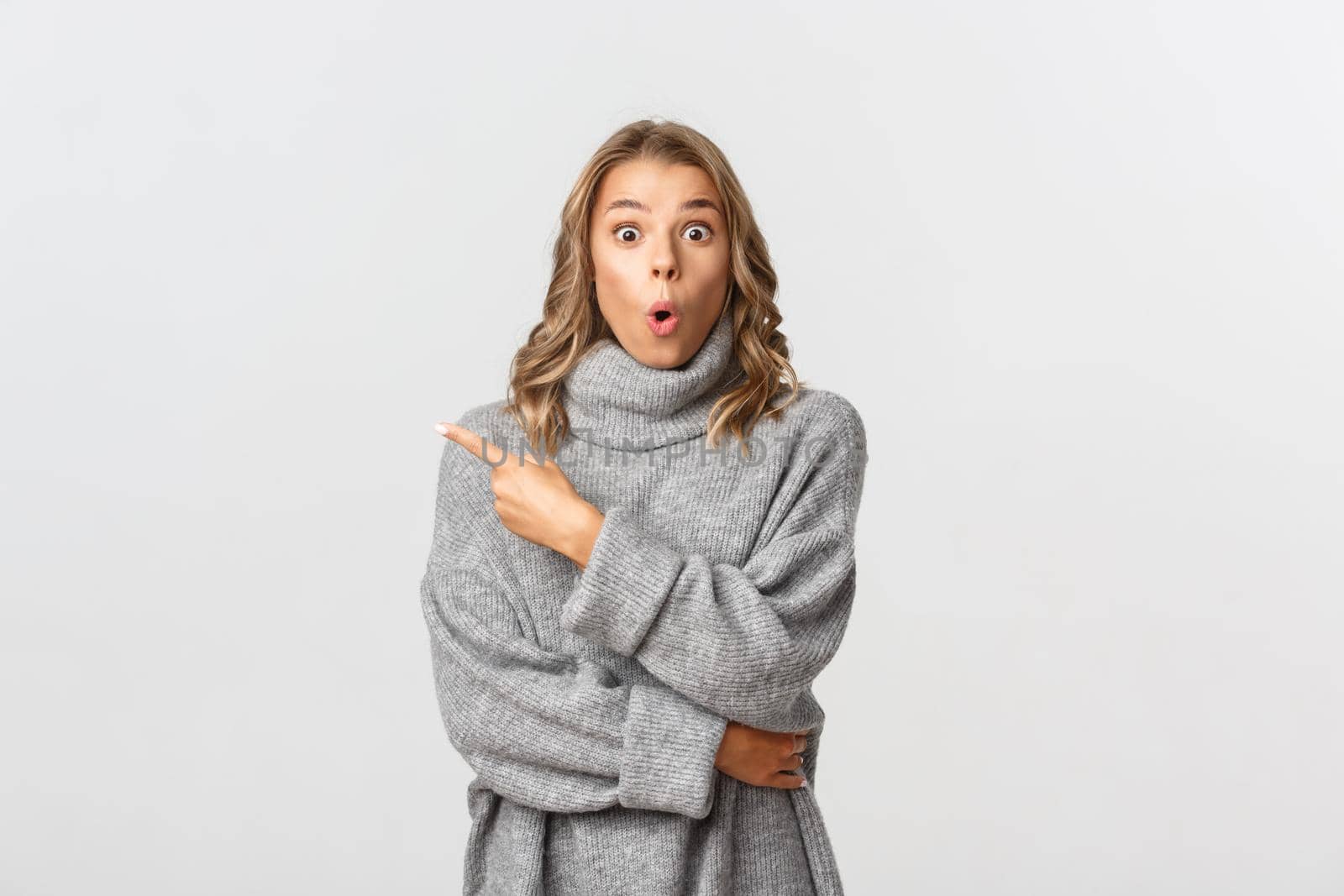 Image of beautiful blond girl in grey sweater pointing finger at upper left corner, showing your logo or banner, looking surprised, standing over white background.