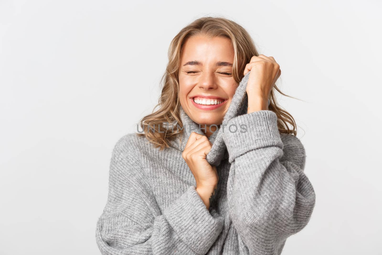 Image of happy attractive girl with blond short hairstyle, touching her soft grey sweater and smiling, standing over white background.