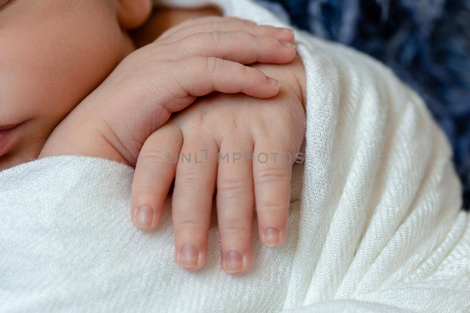 Close-up of little baby handles. The sleeping Newborn boy under a white knitted blanket lies on the blue fur. Newborn. 14 days.
