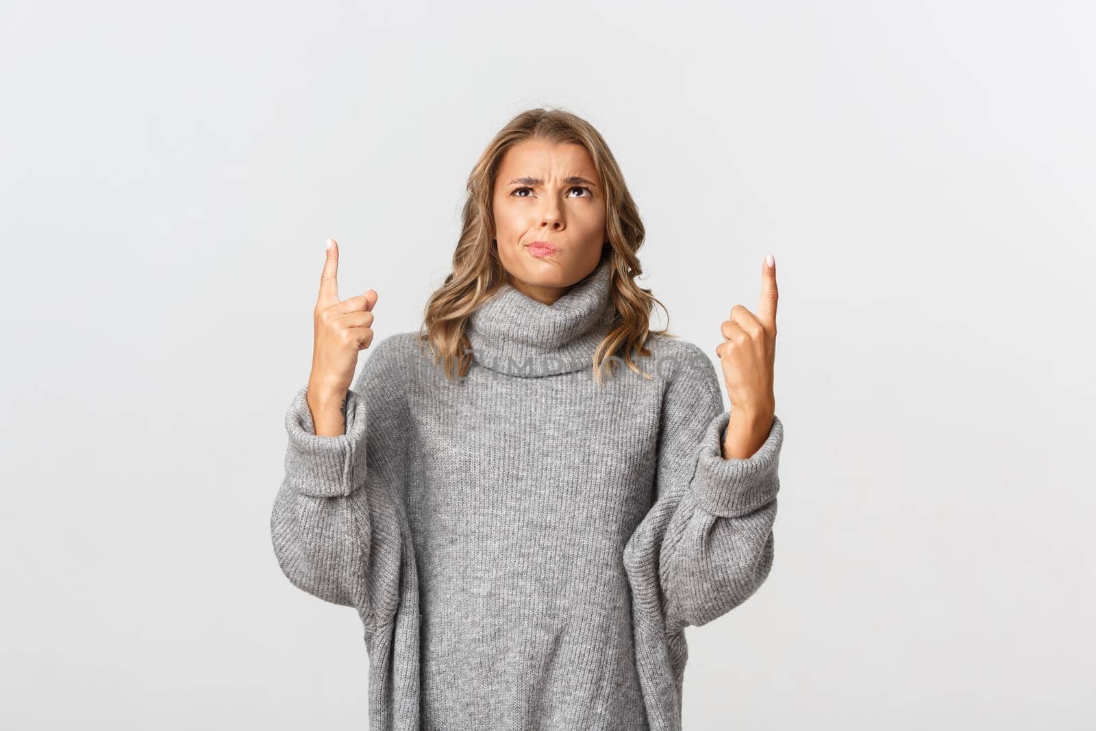 Image of skeptical blond girl in grey sweater, looking indecisive and pointing up, smirking doubtful, standing over white background by Benzoix