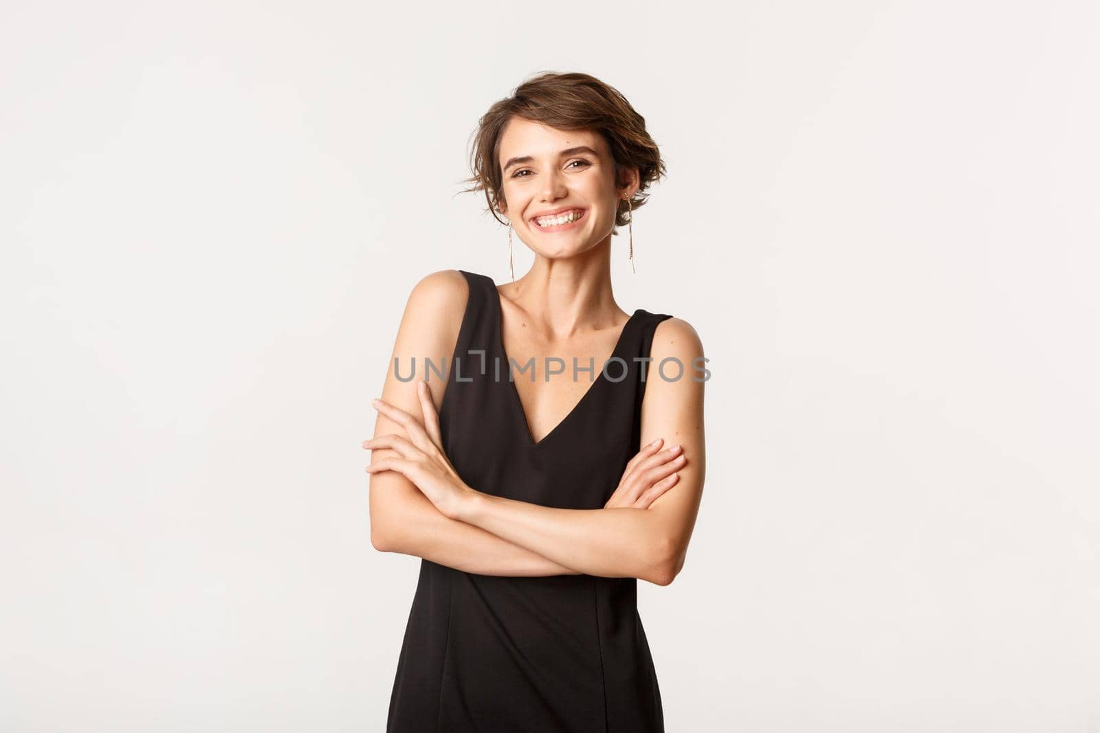 Image of successful attractive young woman in elegant black dress, cross arms chest confident and smiling happy, standing over white background.