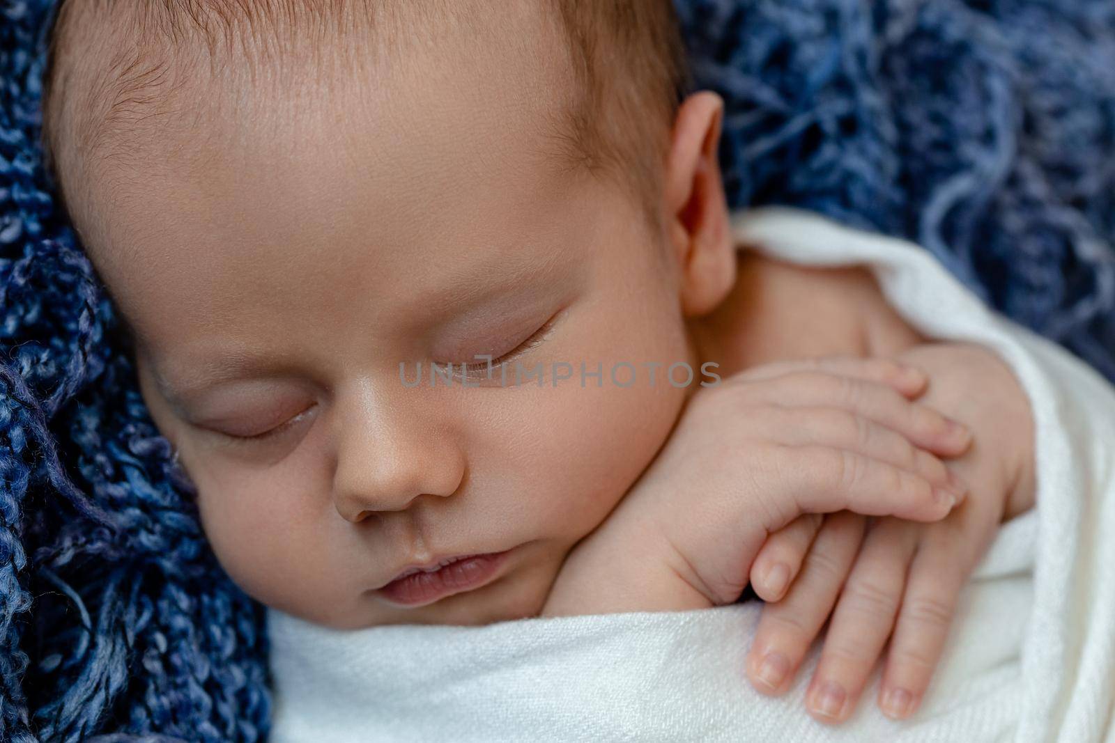 Newborn - baby, face close-up. The sleeping Newborn boy under a white knitted blanket lies on the blue fur. Newborn. 14 days.