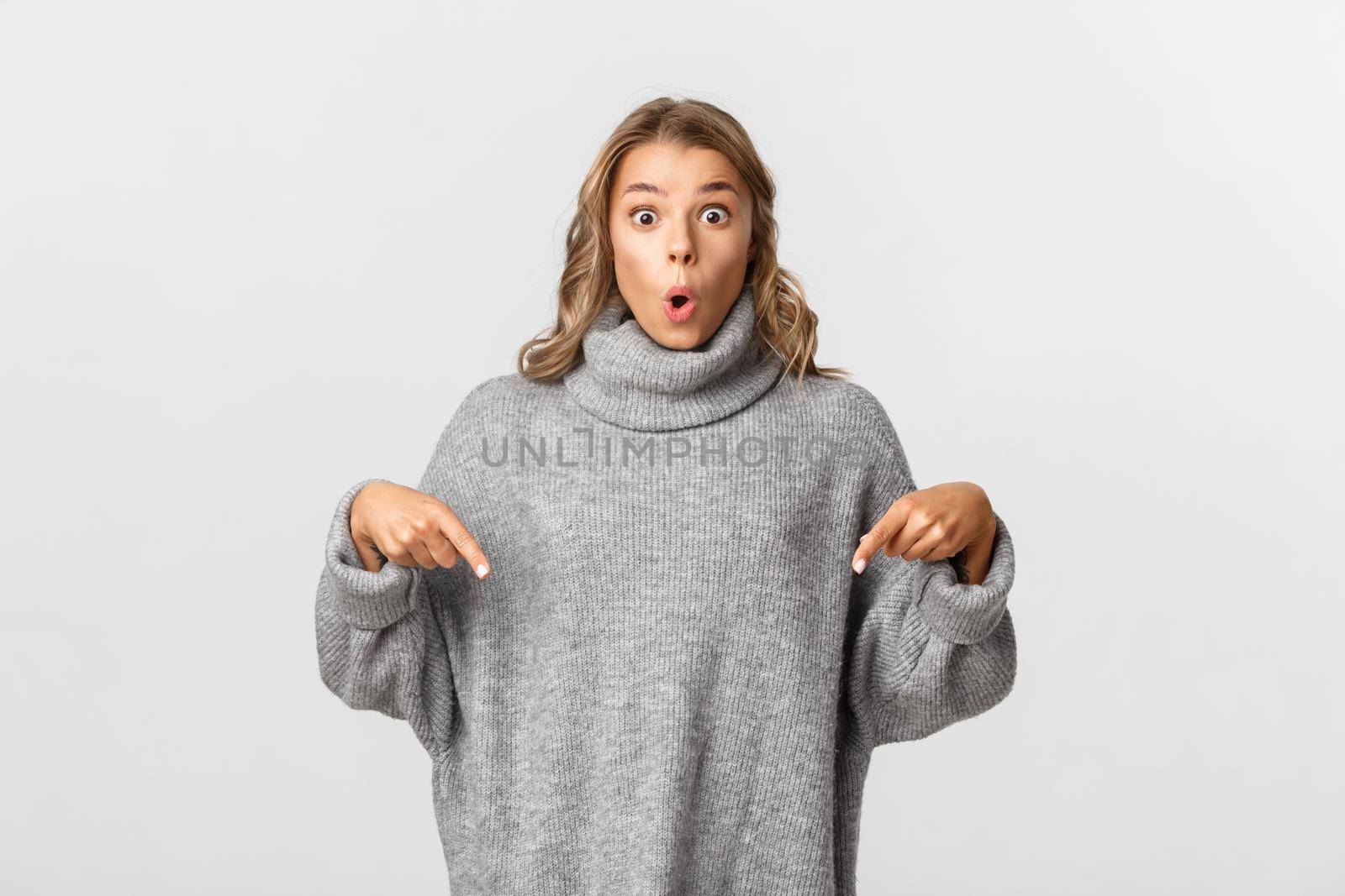 Portrait of surprised girl in grey sweater pointing fingers down, looking impressed with product or brand, white background.