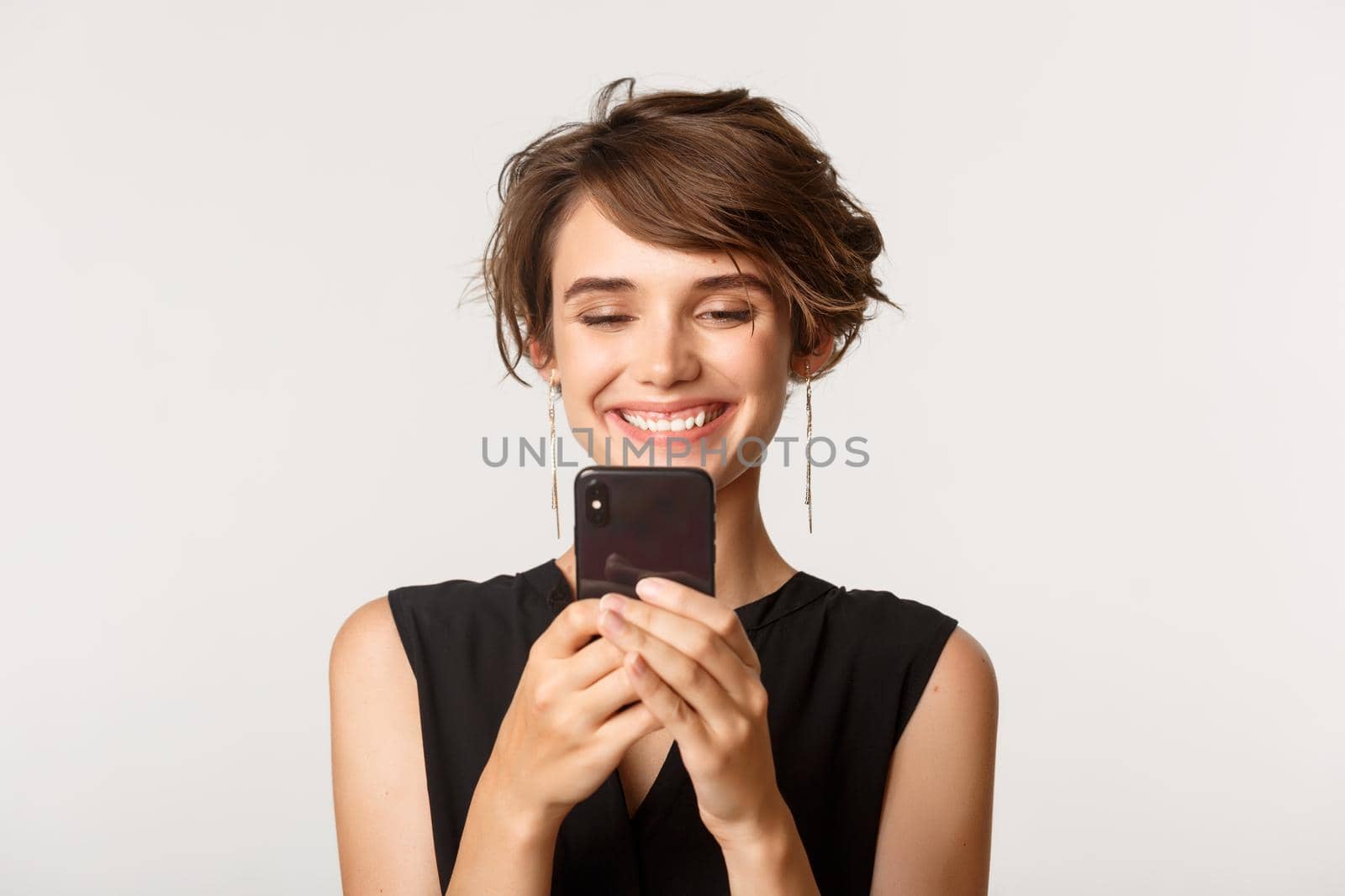 Close-up of satisfied young woman looking at mobile phone with pleased smile, standing over white background by Benzoix